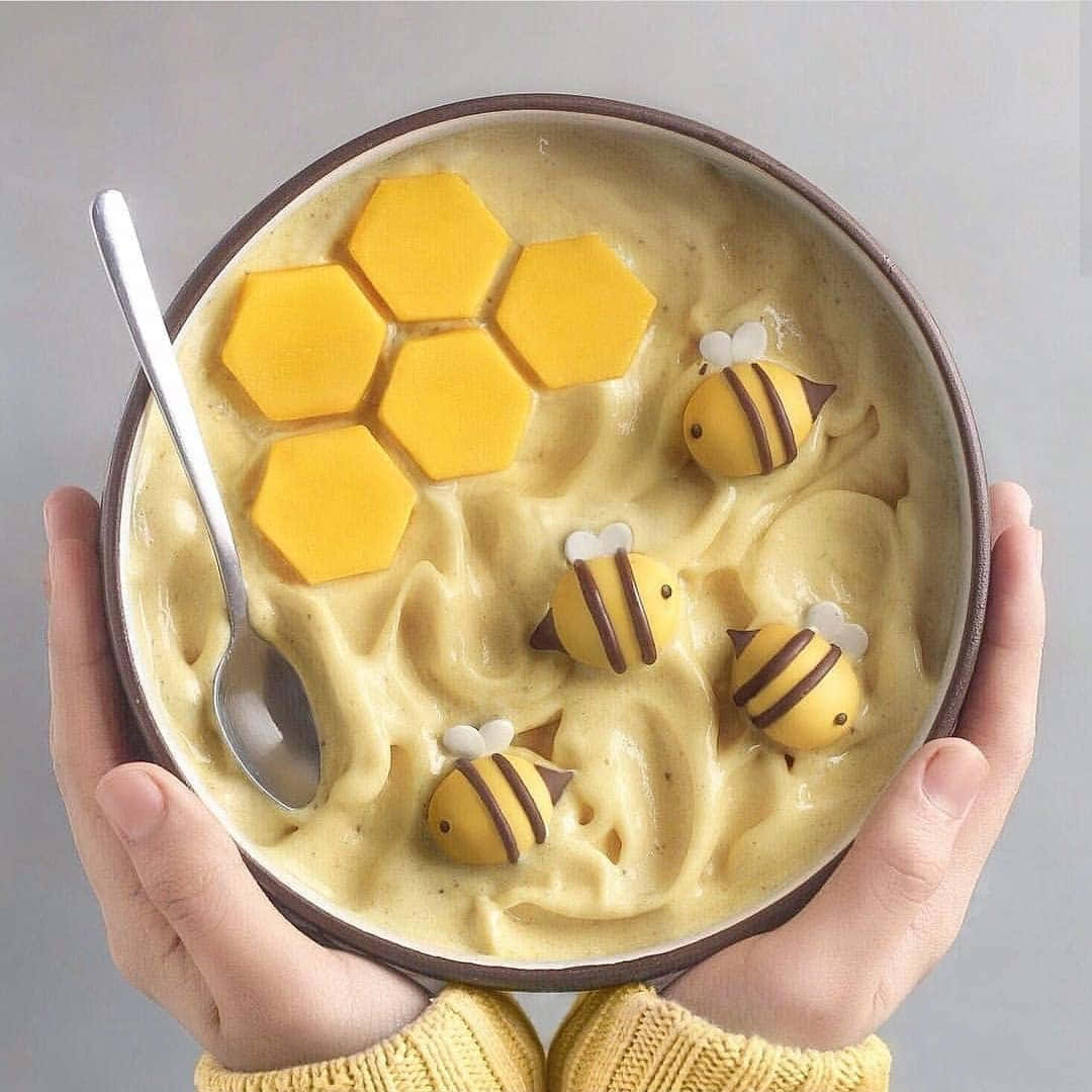 黃色蜜蜂食物美學圖 壁紙 — 雙手捧著一碗奶油黃色甜品，上面放有蜂巢片和可愛的蜜蜂裝飾。
