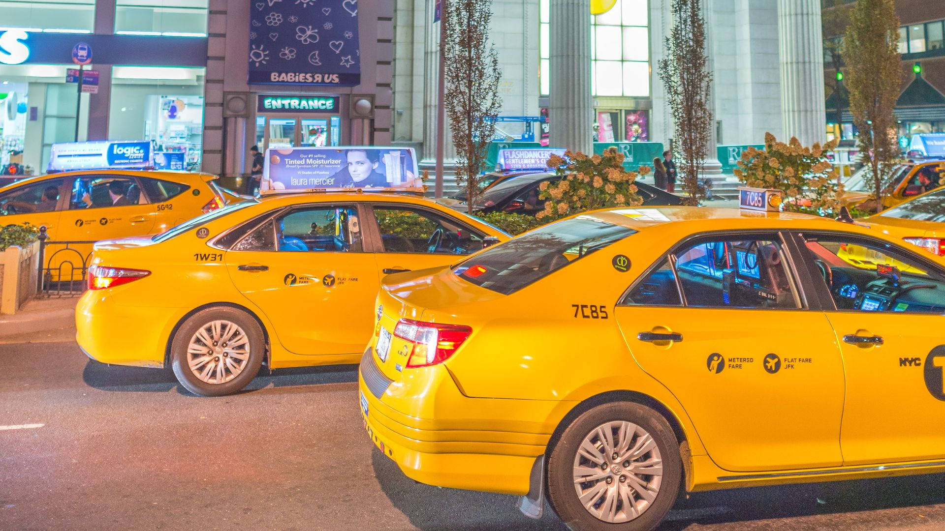黃色計程車停在商場前 壁紙 — 鮮黃色的紐約的士在夜晚排列在繁忙的城市街道上，伴隨著發光的商店櫥窗和行人。