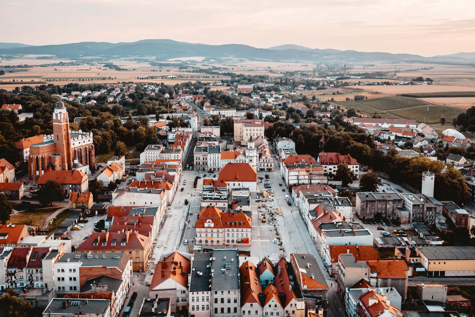 Poland 桌布 壁紙 — 黃金時刻，俯瞰一座迷人的歐洲小鎮，紅瓦屋頂和一座歷史悠久的教堂坐落在起伏的丘陵之間。