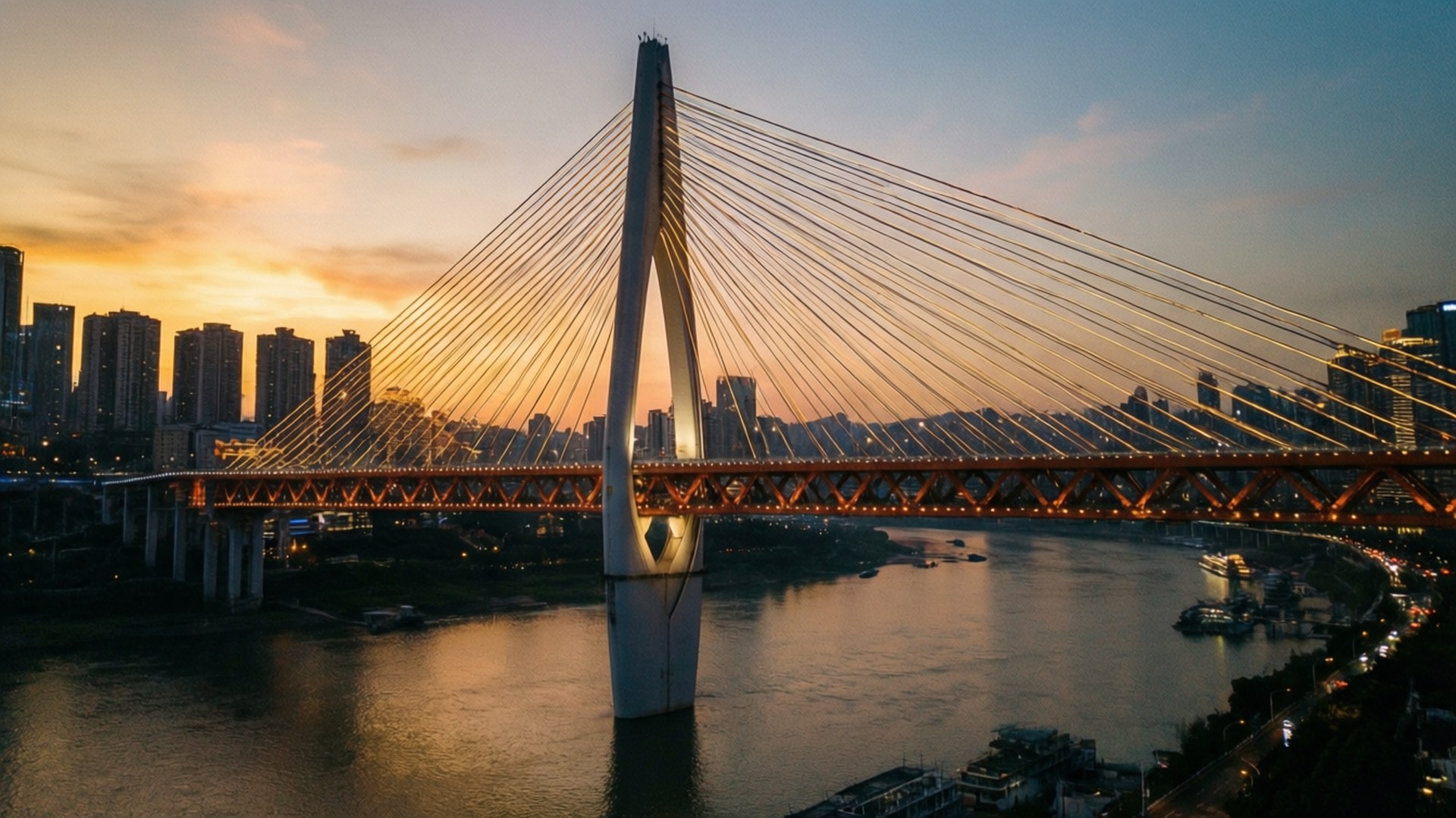 一座亮燈的斜拉橋在日落時分橫跨河流，後面是城市建築。