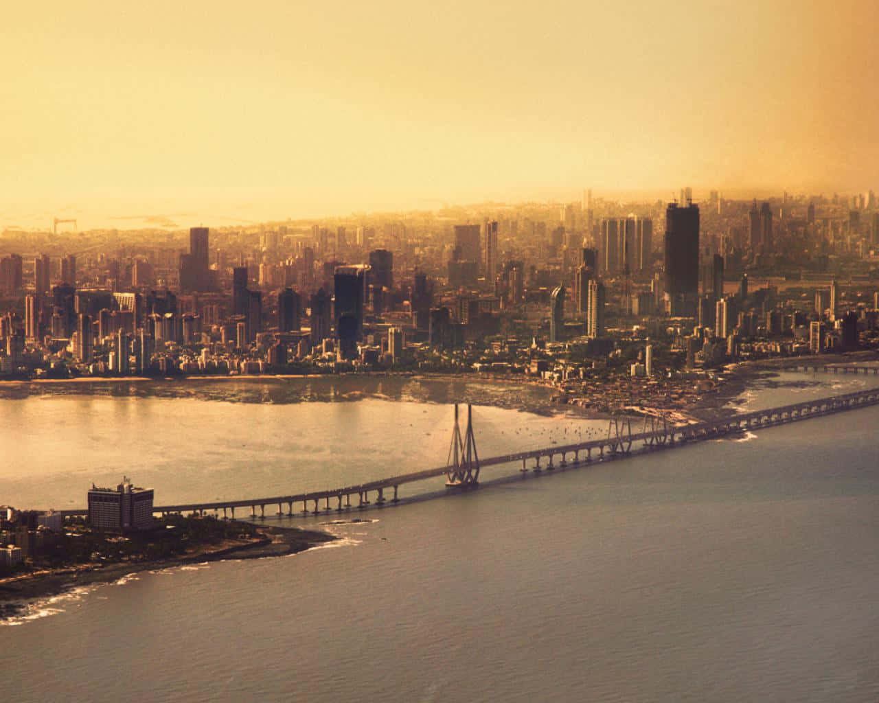 Mumbai 桌布 壁紙 — 黃金時段的光芒沐浴著廣闊的海岸城市景觀，一座長橋橫跨平靜的水面，延伸向摩天大樓。