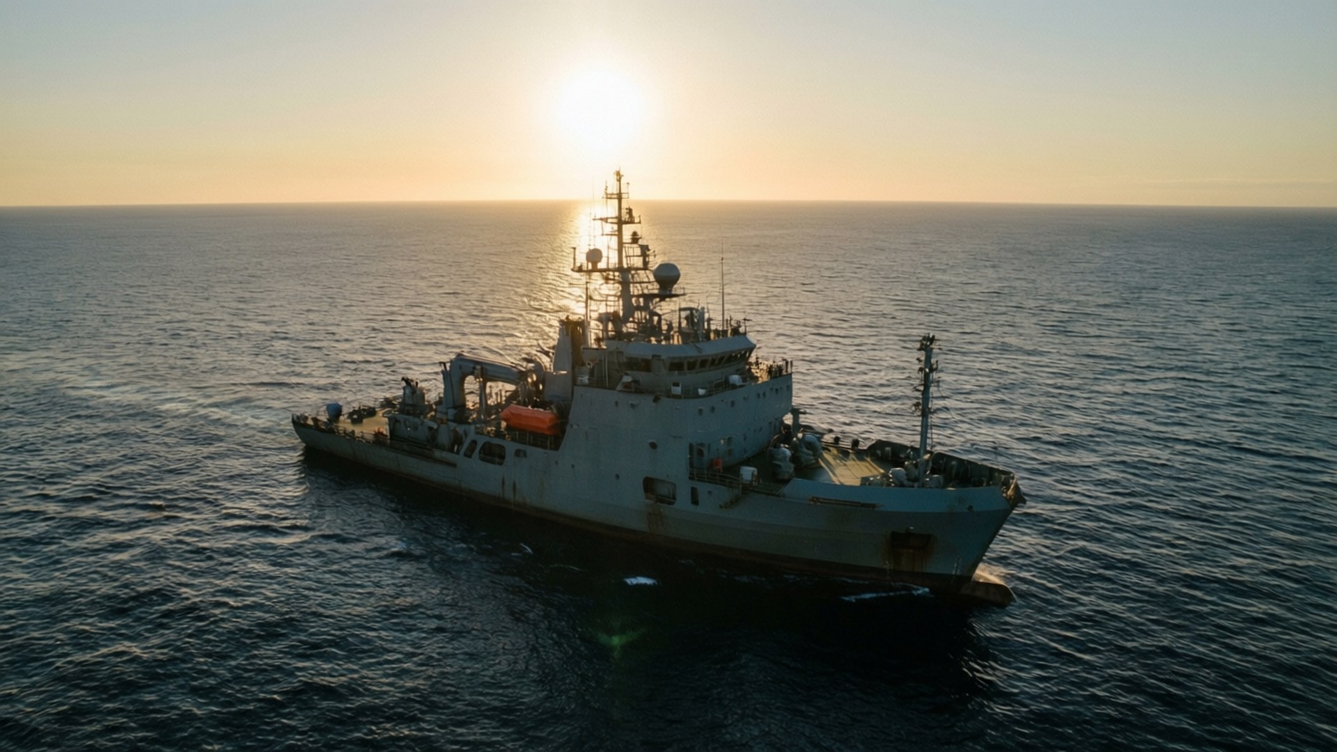 黎明搜尋 壁紙 — 一艘大型軍艦在開闊的海洋上平穩航行，夕陽在它身後落下，將金色光芒灑在水面上。