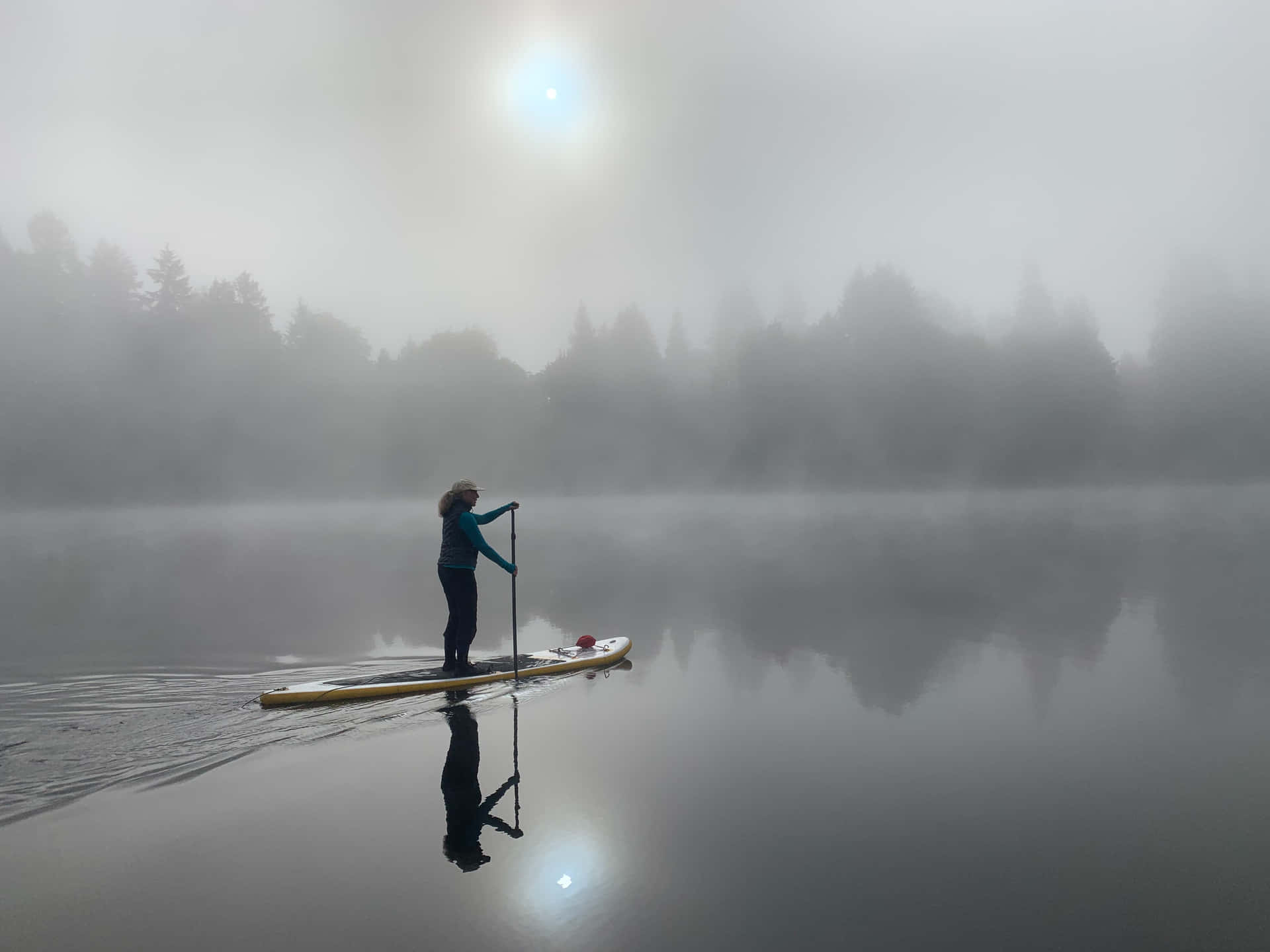 Paddleboarding 桌布 壁紙 — 黎明時分，一位孤獨的槳板划手在霧氣瀰漫的湖面上滑行，樹木逐漸消失在霧中，上方陽光刺眼。