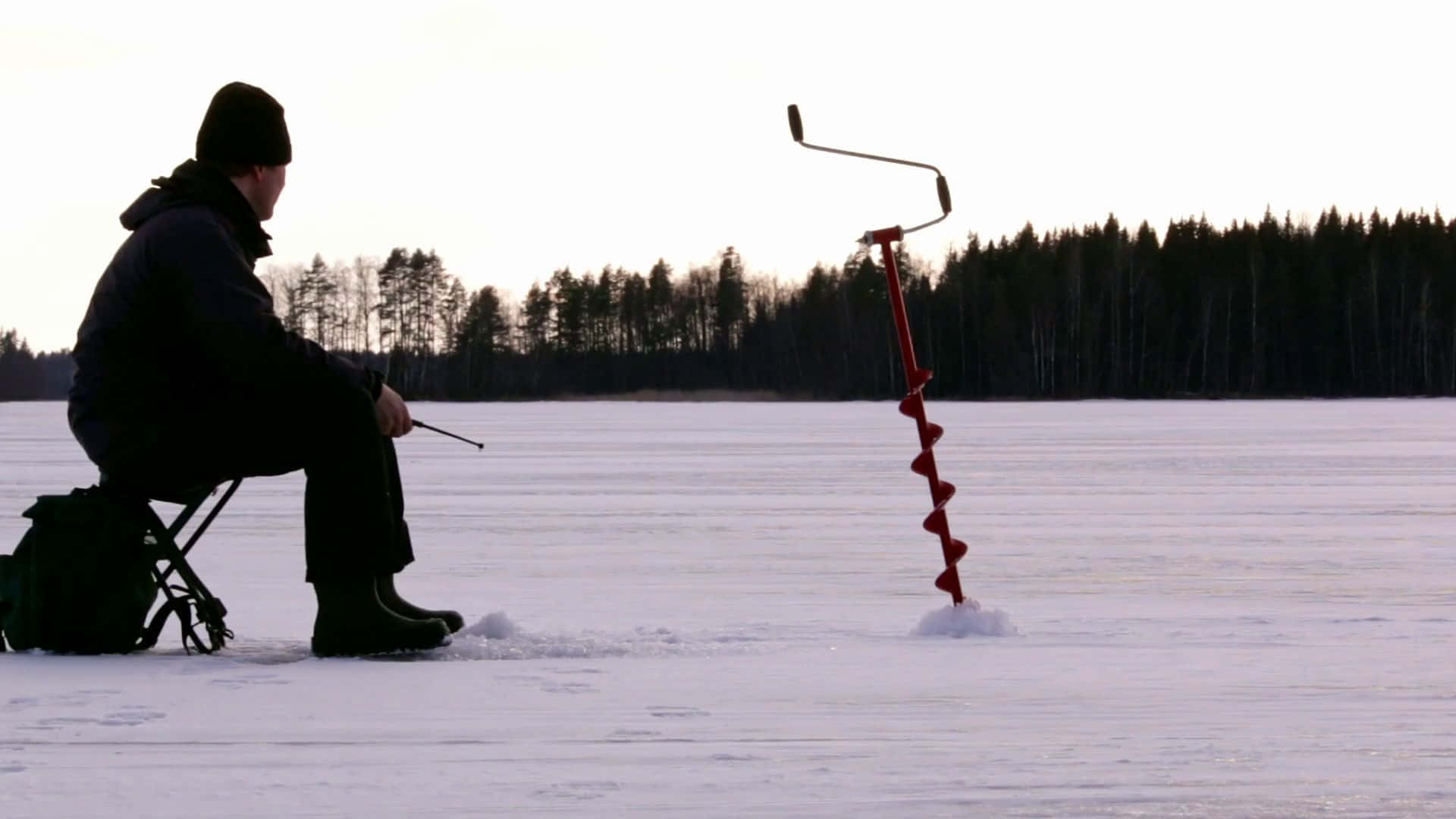 Ice Fishing 桌布 壁紙 — 黎明時分，一名孤獨的漁夫坐在冰凍湖面上的凳子上，用紅色冰鑽鑽洞。