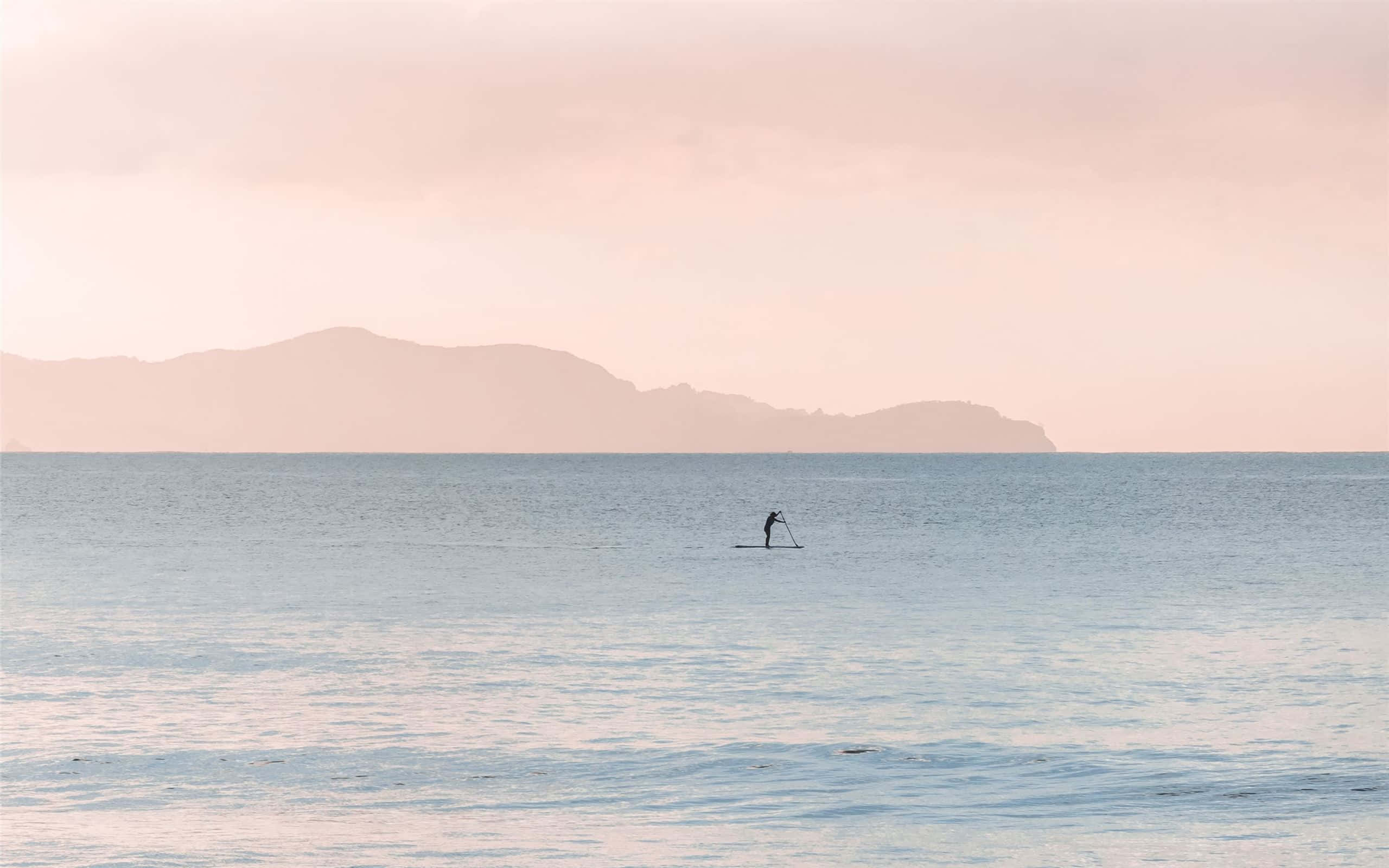 Paddleboarding 桌布 壁紙 — 黎明時分，一名獨自划槳板的人在平靜的海面上滑行，柔和的粉紅色和藍色色調反射在水面和遠處朦朧的山巒上。
