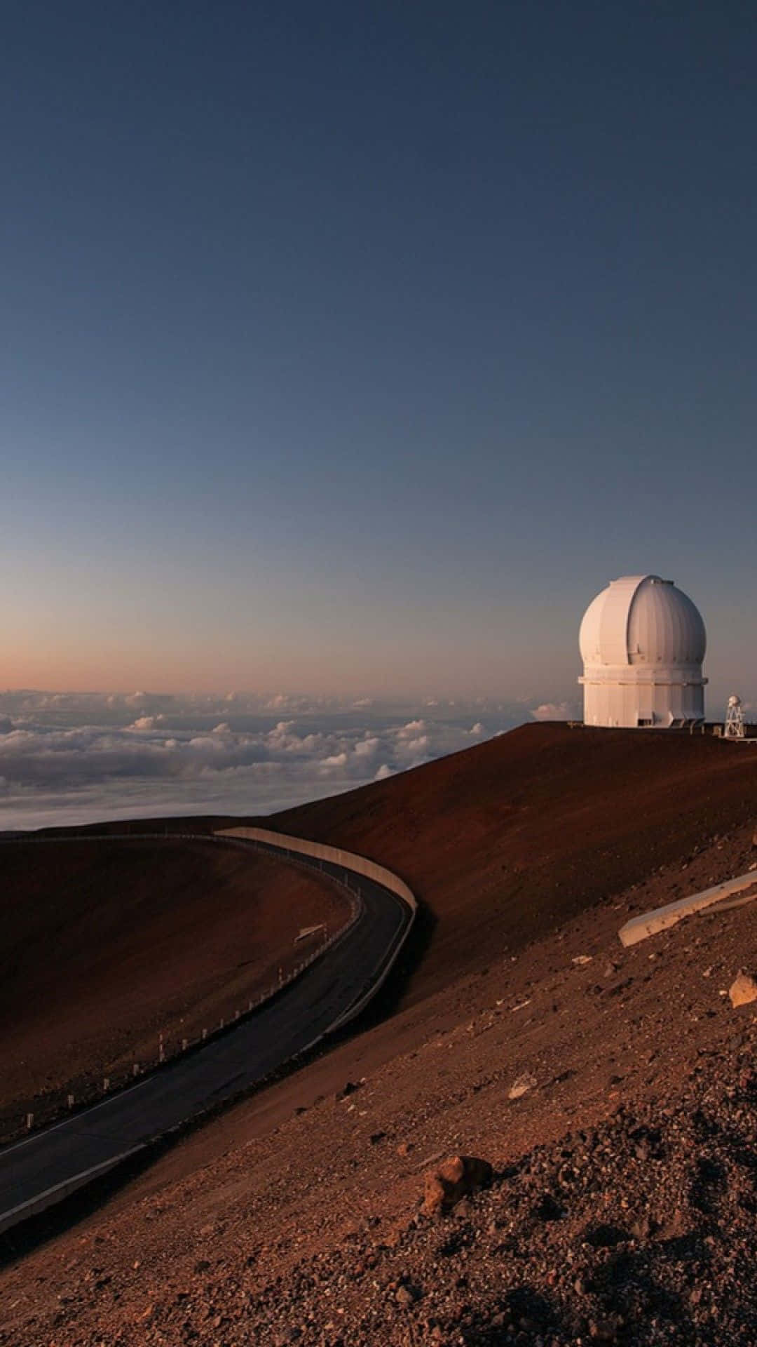 Space Observation 桌布 壁紙 — 黎明時分，一座白色天文台圓頂矗立在崎嶇的火山山頂上，下方是蜿蜒的道路，雲層一直延伸到地平線。