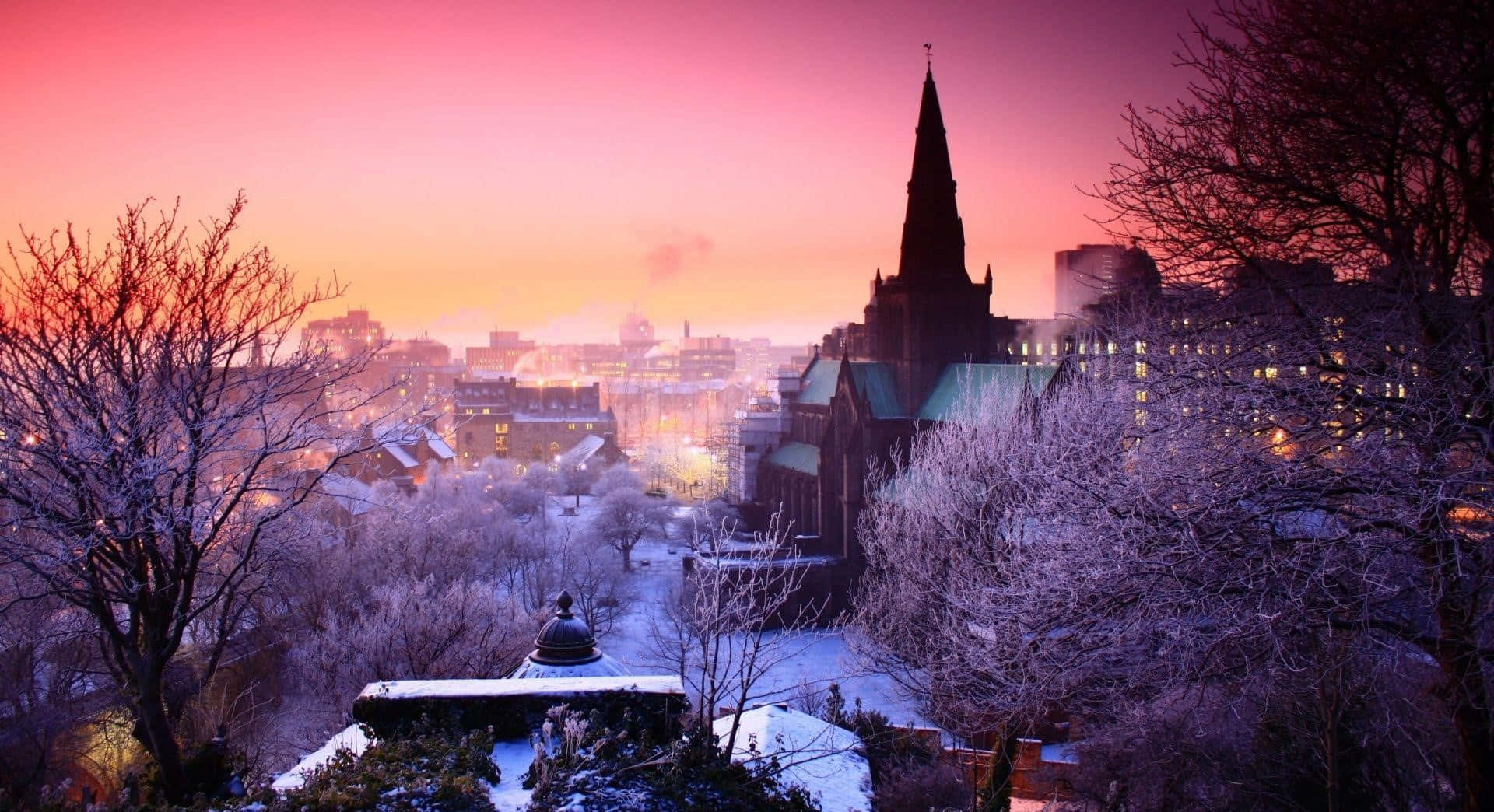Winter Cityscape 桌布 壁紙 — 黎明時分，粉紅色和紫色天空下，白雪覆蓋的城市景觀，帶有哥德式教堂尖頂和結霜的樹木。