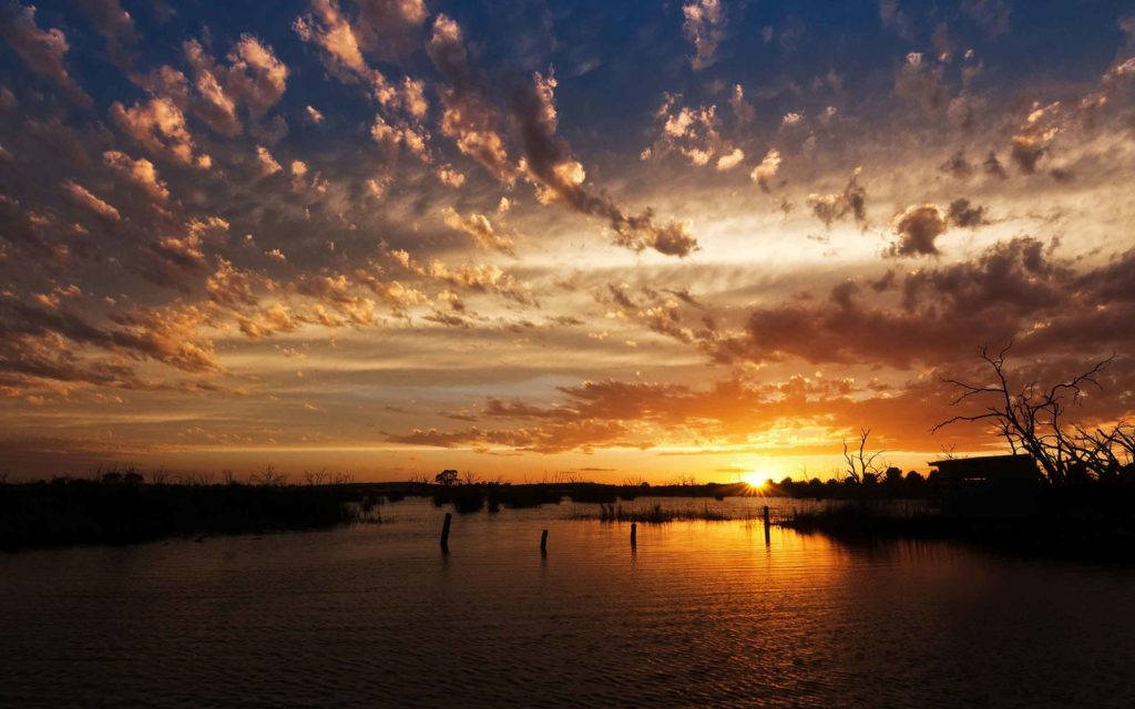 黎明時分湖畔日出自然風景 壁紙 — 充滿活力的日落將天空染成火熱的橙色和深藍色，雲彩倒映在平靜的水面上，樹木和柱子的剪影清晰可見。