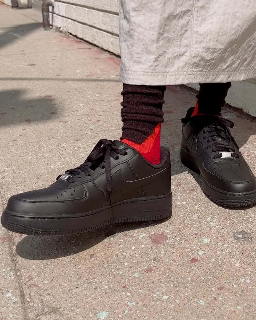 黑人空軍飛行員翱翔天際 壁紙 — 黑色Nike Air Force 1波鞋特寫，露出鮮紅色襪子，穿著在陽光普照的混凝土行人路上。