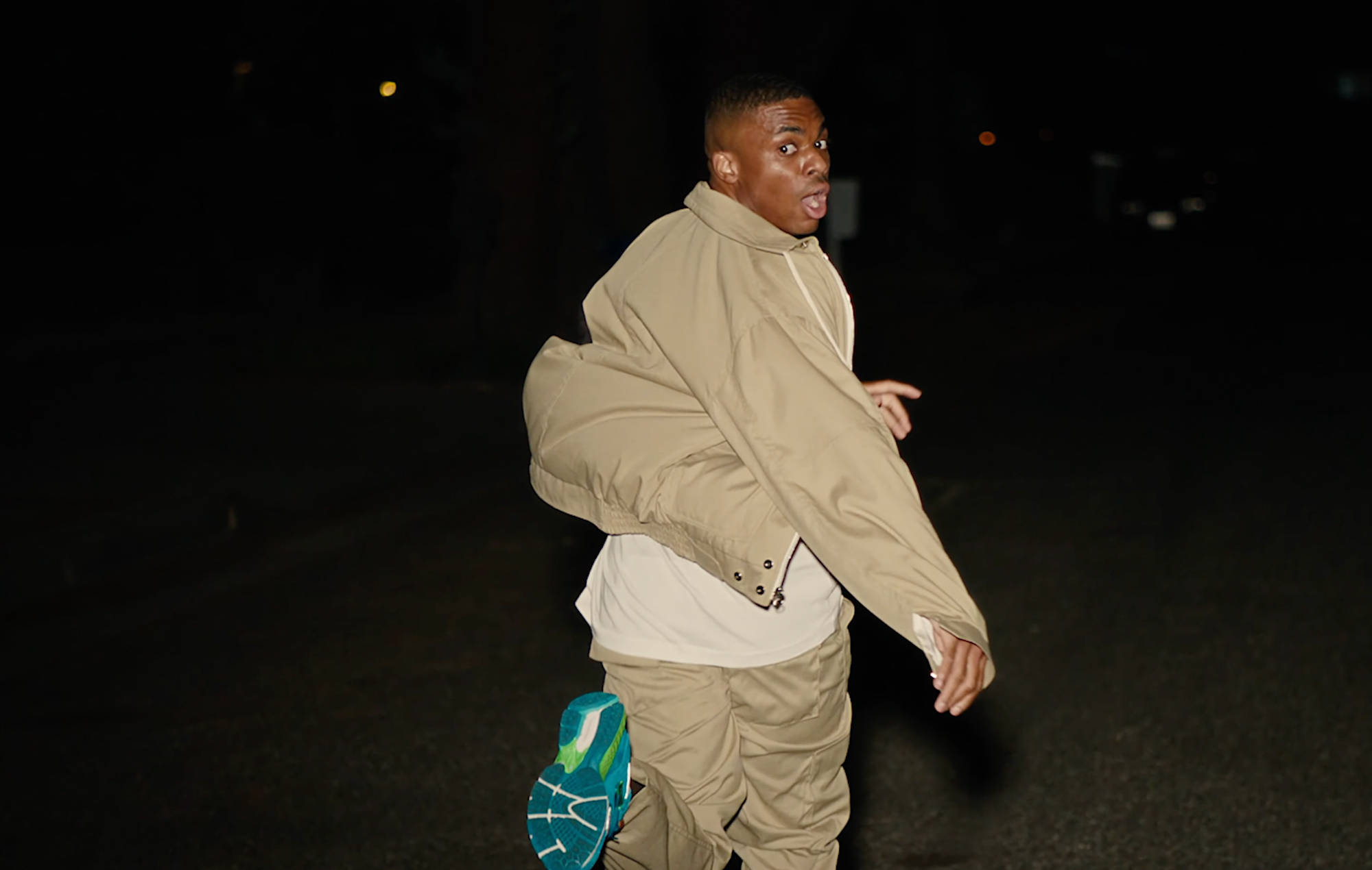 黑人饒舌歌手 Vince Staples 壁紙 — 一名身穿米色外套和褲子的年輕男子在夜晚的黑暗街道上奔跑，可見一隻亮藍色的鞋子。