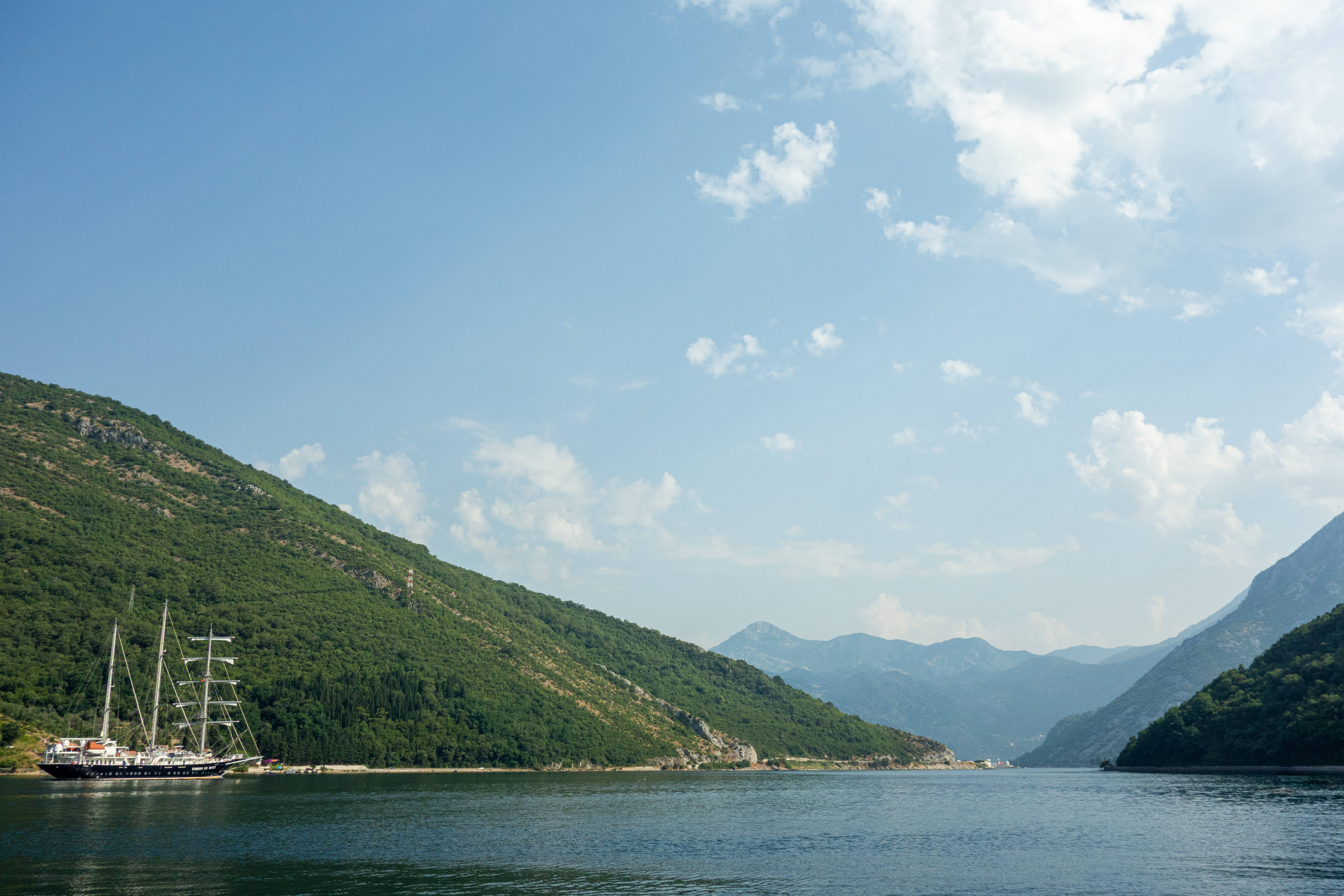 黑山卡梅納里風景 壁紙 — 一艘大型帆船在深藍色峽灣中寧靜地停泊，兩側是鬱鬱蔥蔥的綠色山脈，天空明亮，雲朵散佈。