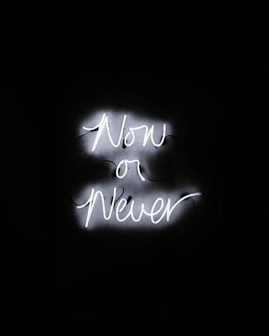 一個發光的白色霓虹燈牌，在黑暗的房間中寫著「Now or Never」。