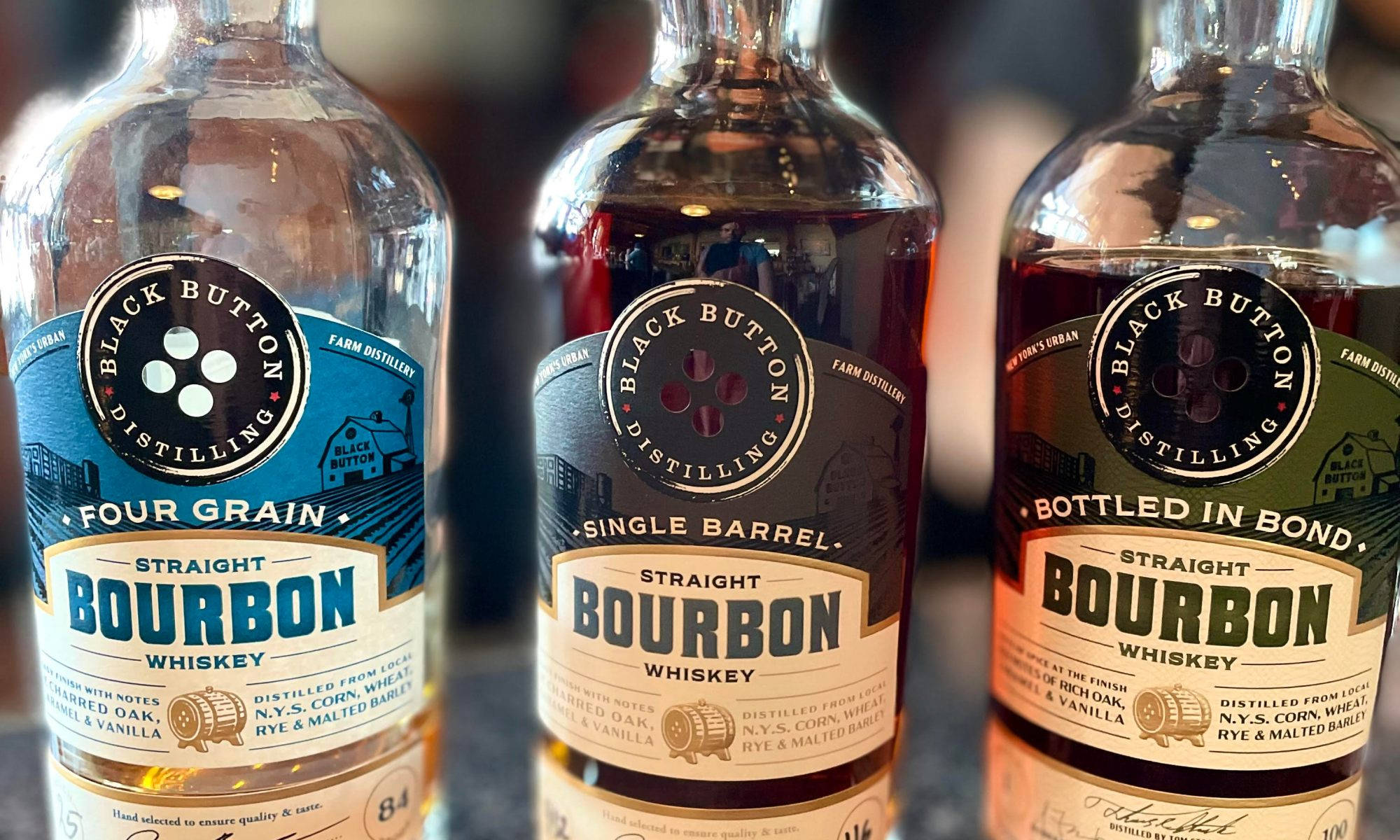 黑按鈕蒸餾廠的手工烈酒 壁紙 — 三瓶 Black Button Distilling 波本威士忌，帶有獨特的標籤和液體顏色，在深色表面上排成一排展示。