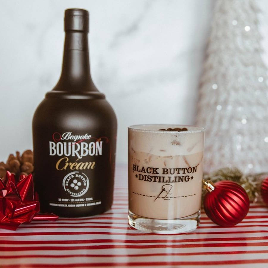黑按鈕蒸餾廠的高級手工烈酒 壁紙 — 一瓶Black Button Bourbon Cream和一杯奶油雞尾酒放在紅色條紋桌布上，旁邊是節日紅色裝飾品和一棵白色聖誕樹。