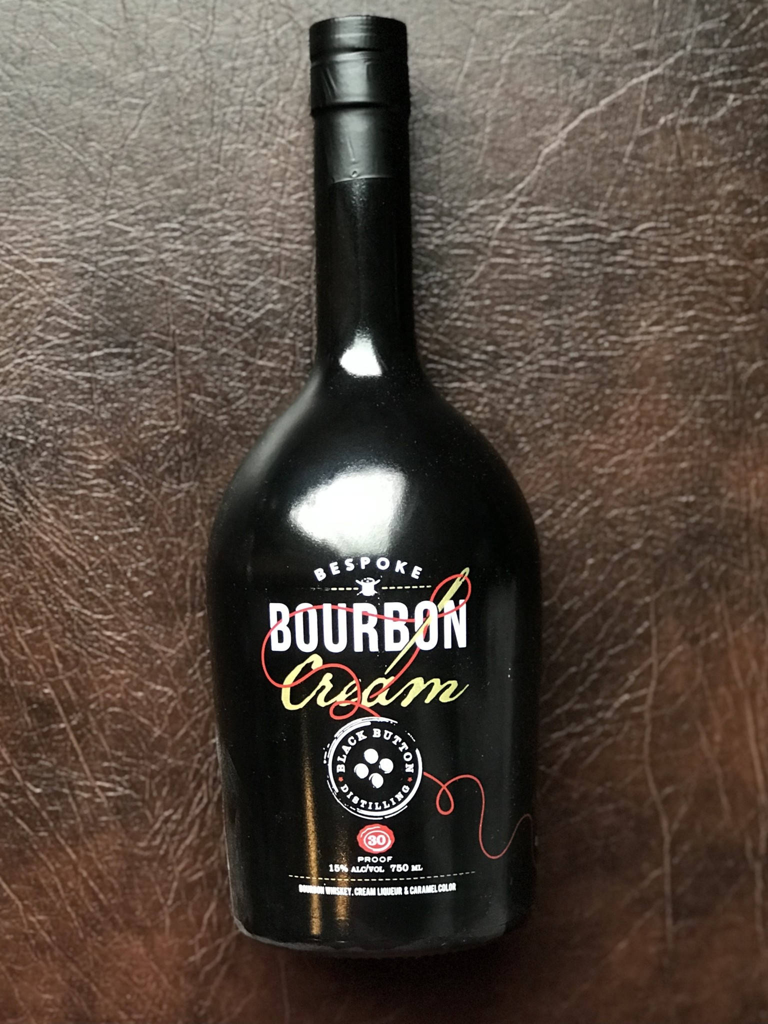 黑按鈕蒸餾廠訂製威士忌 壁紙 — 一個時尚的黑色Bespoke Bourbon Cream利口酒瓶放置在質感豐富的棕色皮革表面上。