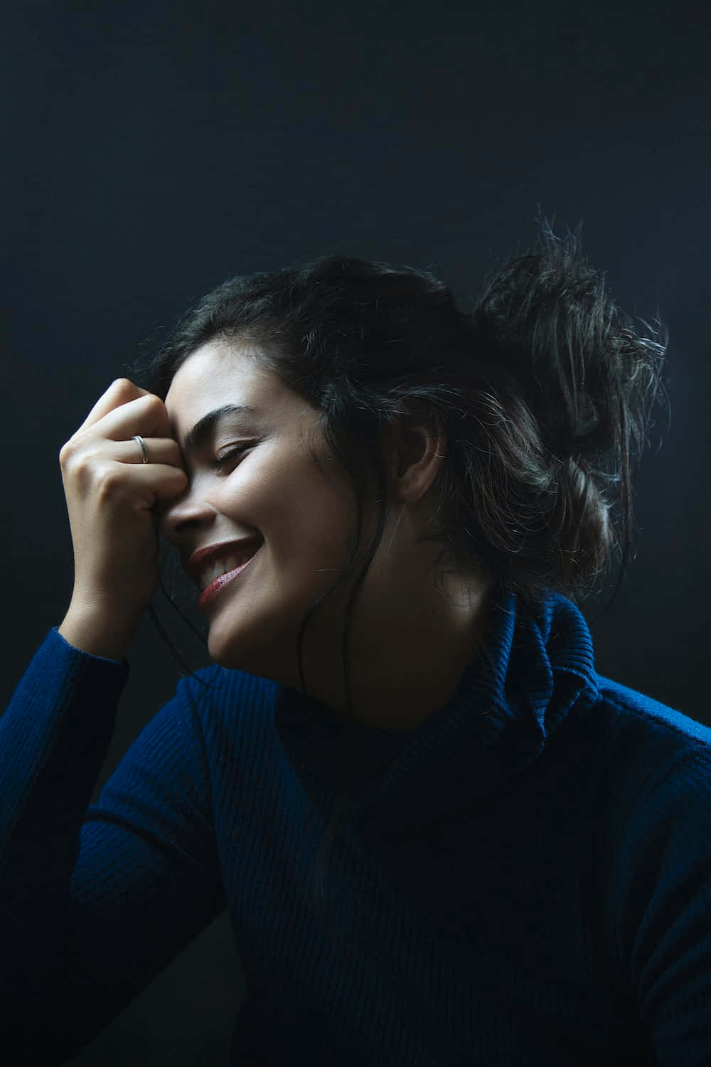一位身穿藍色毛衣的女性，在昏暗的攝影棚中，手靠近眼睛笑著。