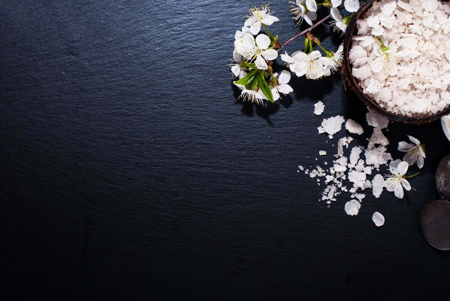 黑木表面的白花水療背景 壁紙 — 白色櫻花和粗鹽從椰子碗中灑落在磨砂黑色石板表面上。