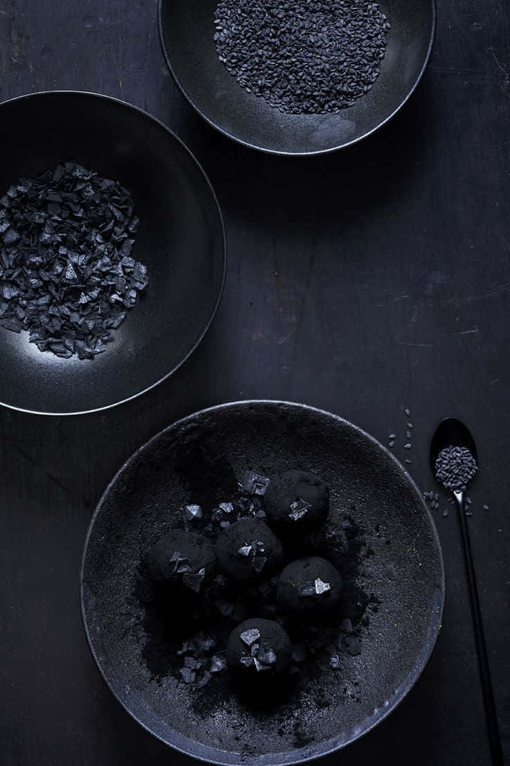 Black Garlic 桌布 壁紙 — 黑朱古力松露配黑芝麻和炭片，擺放在磨砂黑碗中，置於有紋理的表面上。
