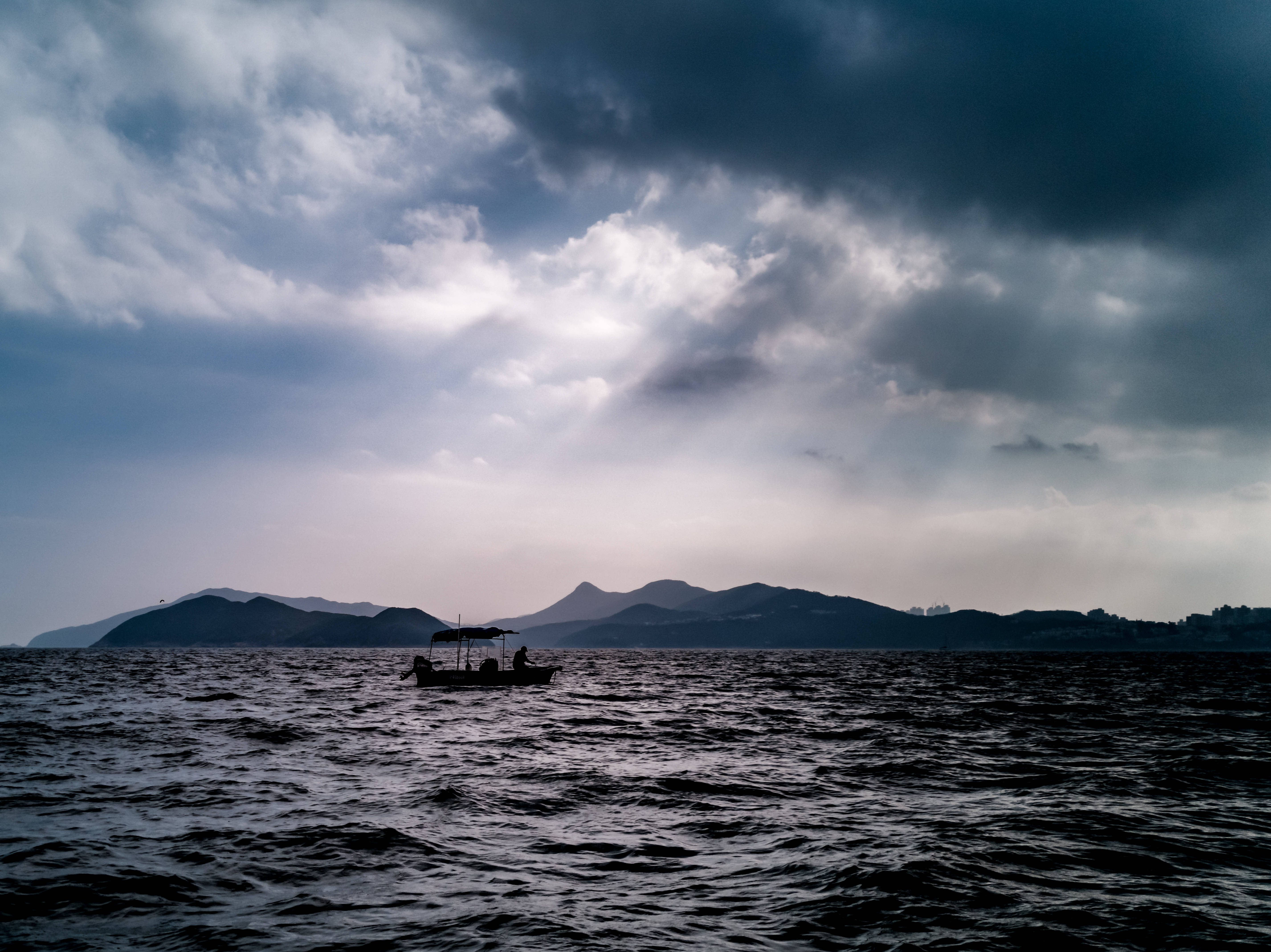 黑海剪影船隻 壁紙 — 一艘孤獨的小船載著兩個人，在波濤洶湧的海面上航行，背景是戲劇性的多雲天空和遠處的山脈。