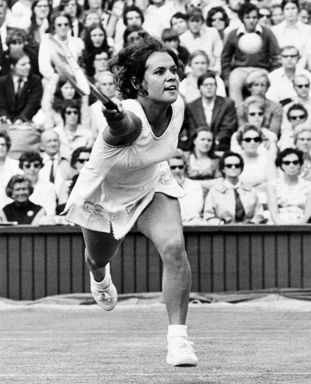黑白 Evonne Goolagong Cawley 網球選手 壁紙 — 一張復古黑白照片，捕捉到一位女網球運動員在草地場上揮拍的中間動作，背景有觀眾。