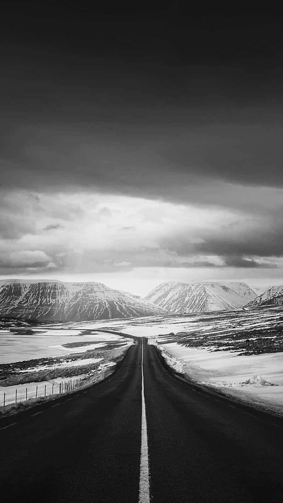 黑白 iPhone 壁紙 — 一條孤獨的道路在戲劇性的單色天空下延伸向積雪覆蓋的山脈。