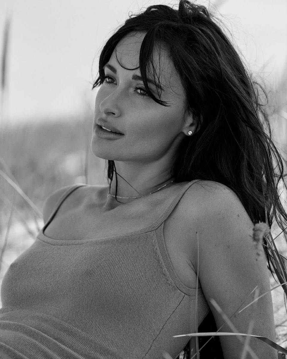 黑白 Kacey Musgraves 照片 壁紙 — 一位黑髮穿背心的女性在高草叢中若有所思地凝視的黑白肖像。