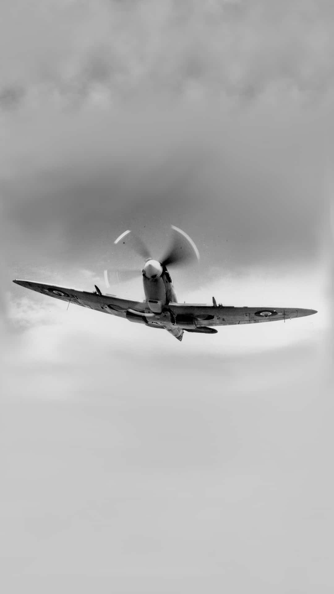 黑白 Supermarine Spitfire Android 飛機背景 壁紙 — 一架復古戰鬥機在陰雲密佈的天空中翱翔，螺旋槳以黑白快速旋轉。