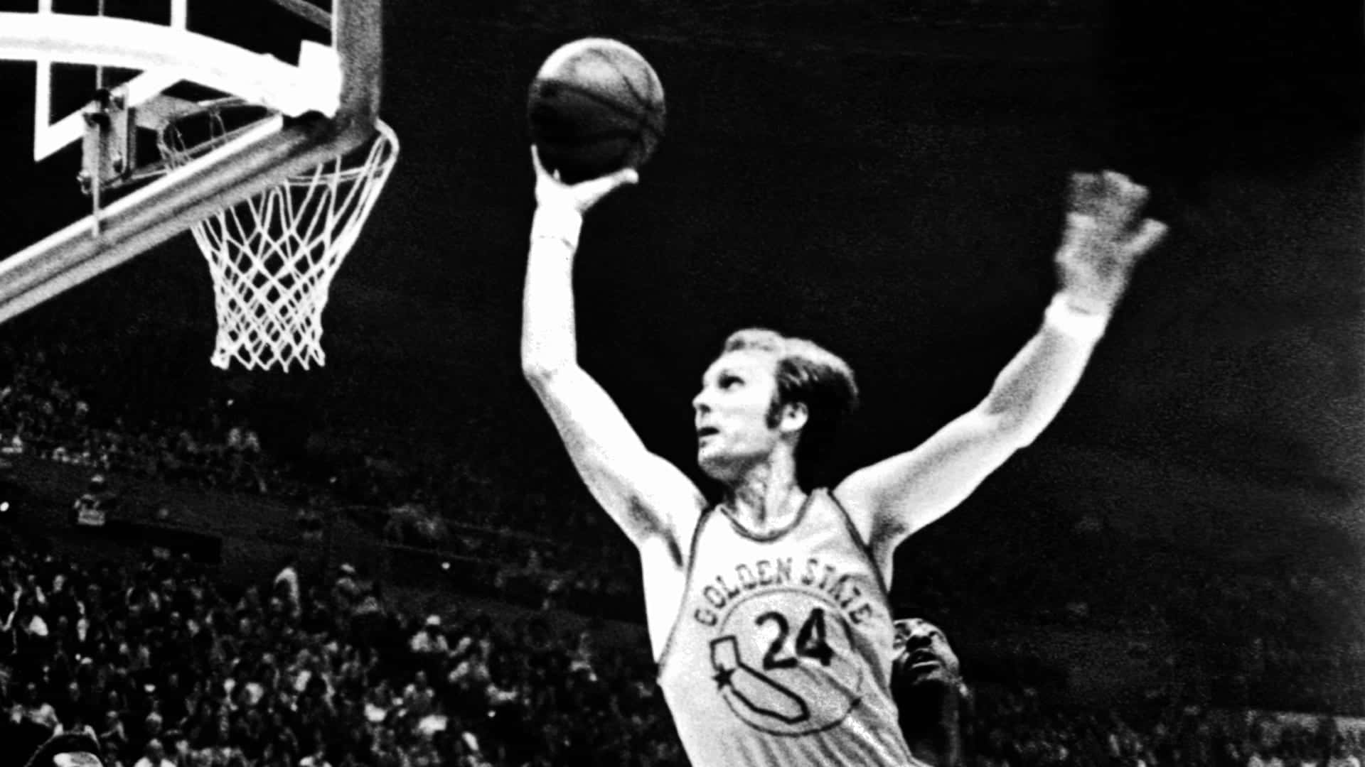 黑白NBA瑞克·巴里灌籃 壁紙 — 一名身穿金州勇士球衣的籃球運動員在明亮的場館燈光下跳起入樽，下方是咆哮的人群。