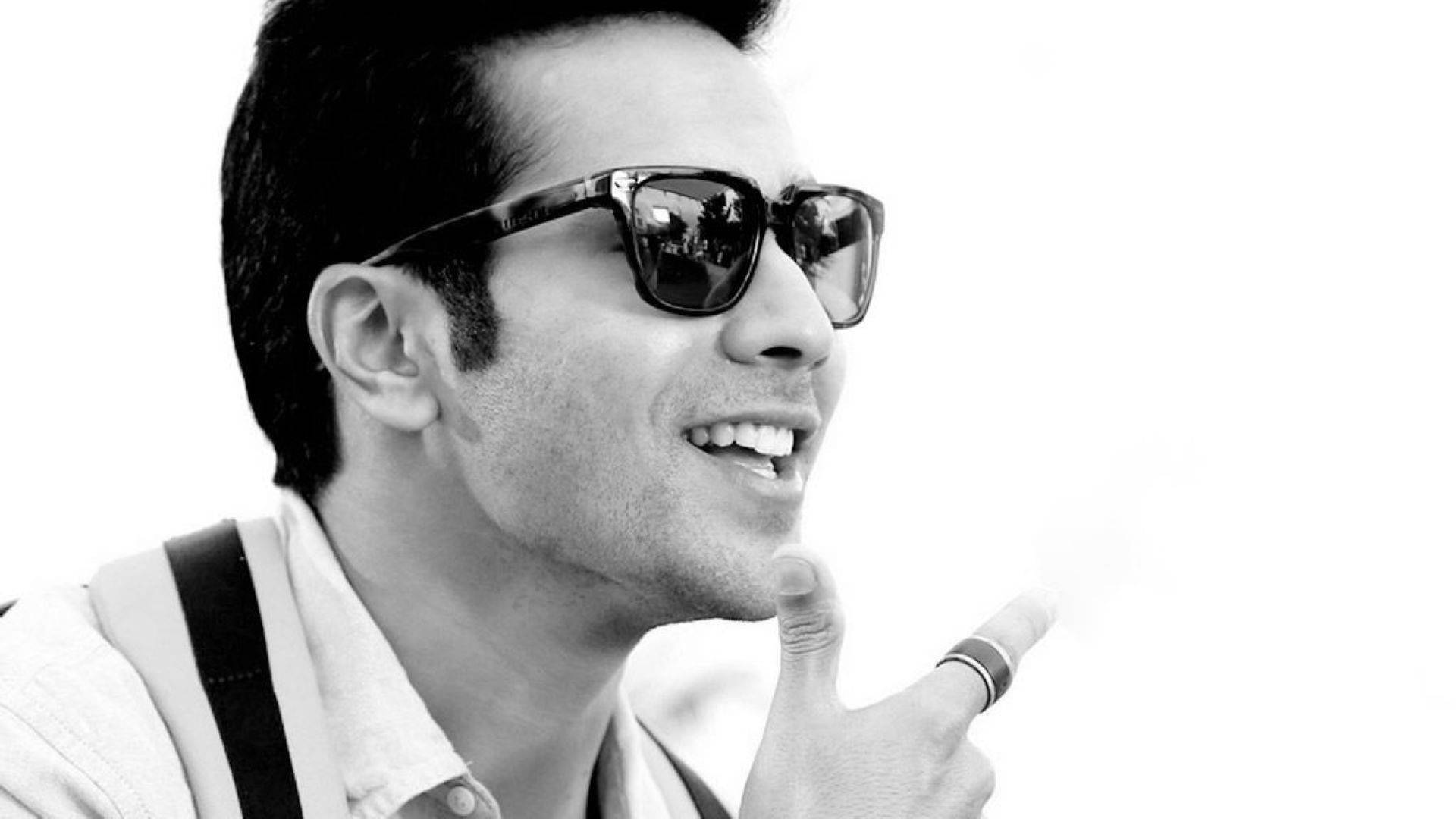 黑白Varun Dhawan 壁紙 — 一位時尚男士戴著太陽眼鏡，在純白背景上拿著香煙微笑。