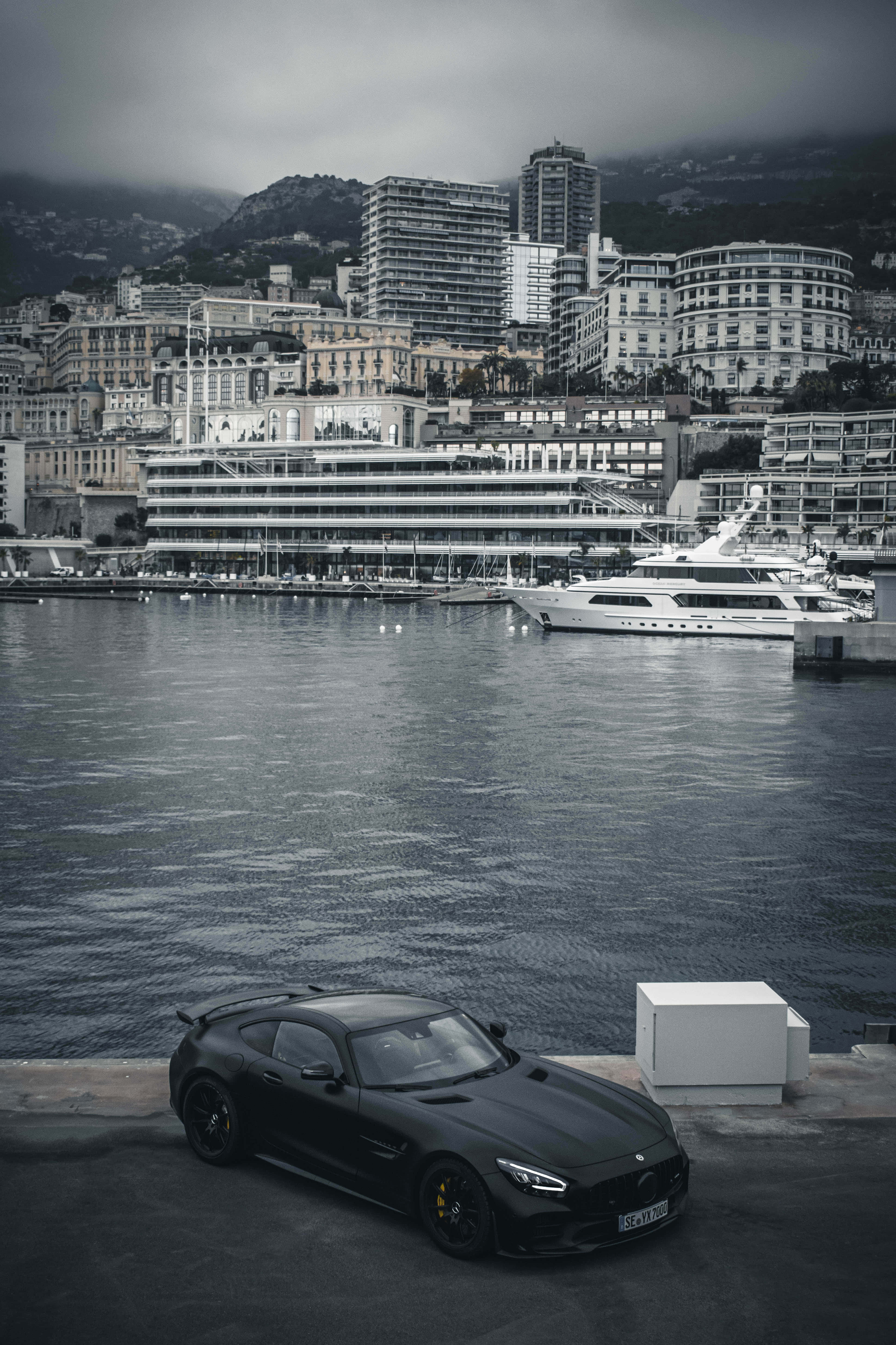 黑白俯瞰安卓AMG GT-R背景 壁紙 — 一架時尚黑色跑車停泊在港口，旁邊有豪華遊艇和懸崖城市，天空多雲。