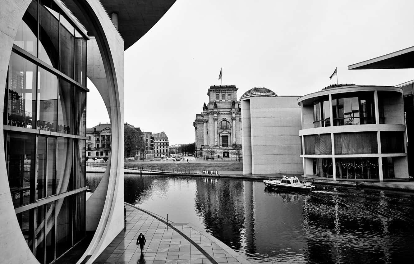 Black And White Architecture 桌布 壁紙 — 黑白城市景觀，現代玻璃建築框住一座歷史建築，橫跨平靜的水道，邊緣有一位孤獨的行人行走。