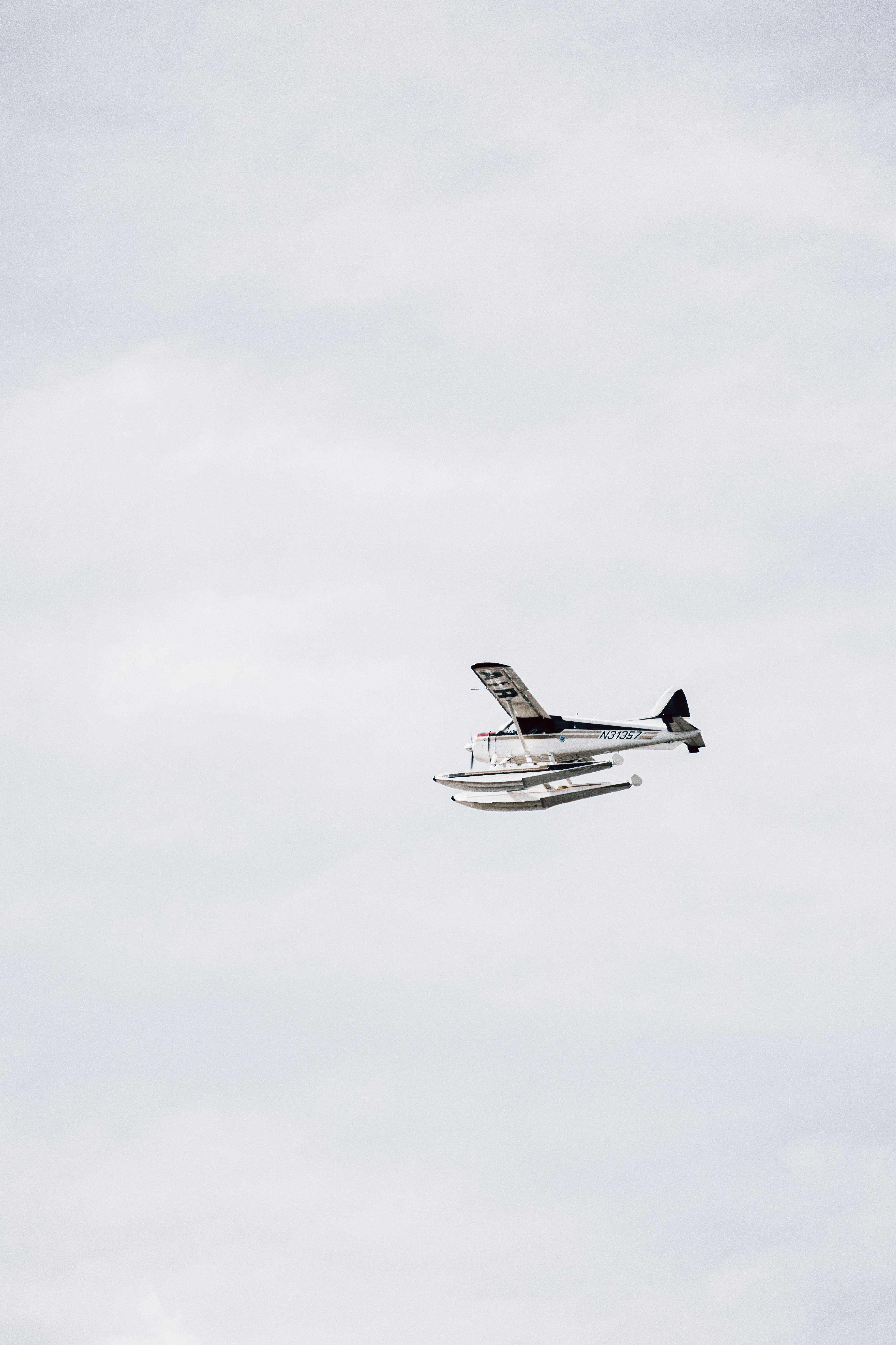 黑白小型飛機起飛 壁紙 — 一架帶有黑色點綴的白色水上飛機在佈滿柔和灰色雲層的廣闊陰天中翱翔。