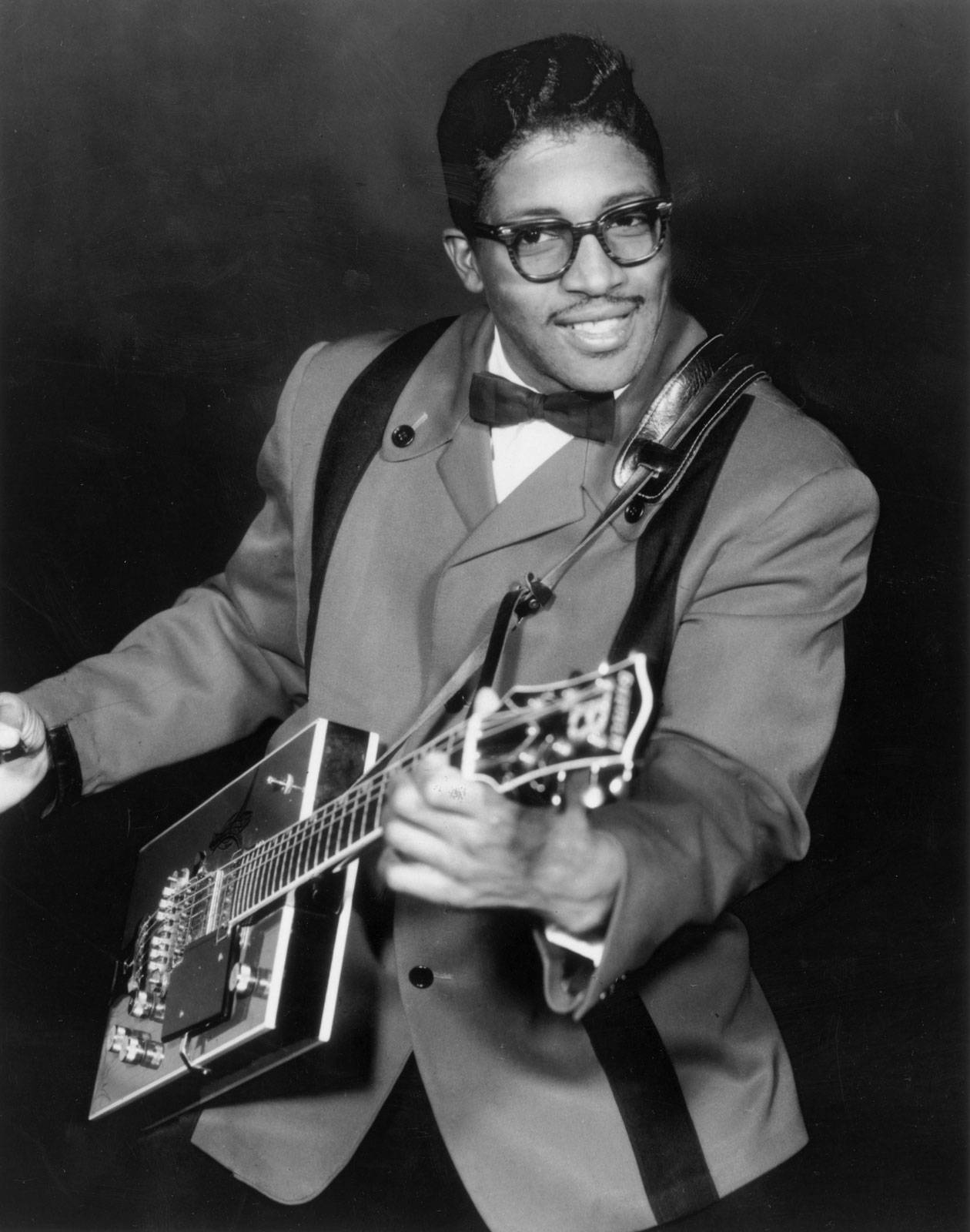黑白年輕Bo Diddley 壁紙 — 一位戴眼鏡、穿西裝的微笑男子，在黑暗的攝影棚背景下拿著一把獨特的電結他。