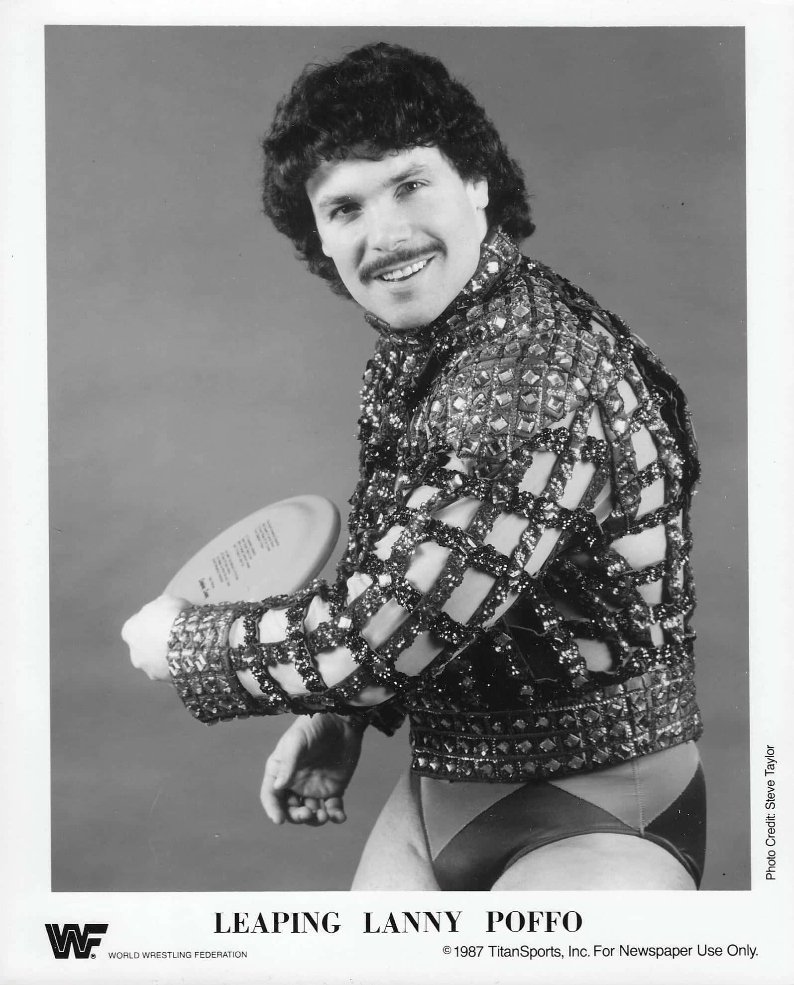 黑白微笑的 Lanny Poffo 壁紙 — 摔角手 Lanny Poffo 穿著閃亮網狀服裝，在簡單的攝影棚背景前拿著飛盤的黑白肖像。