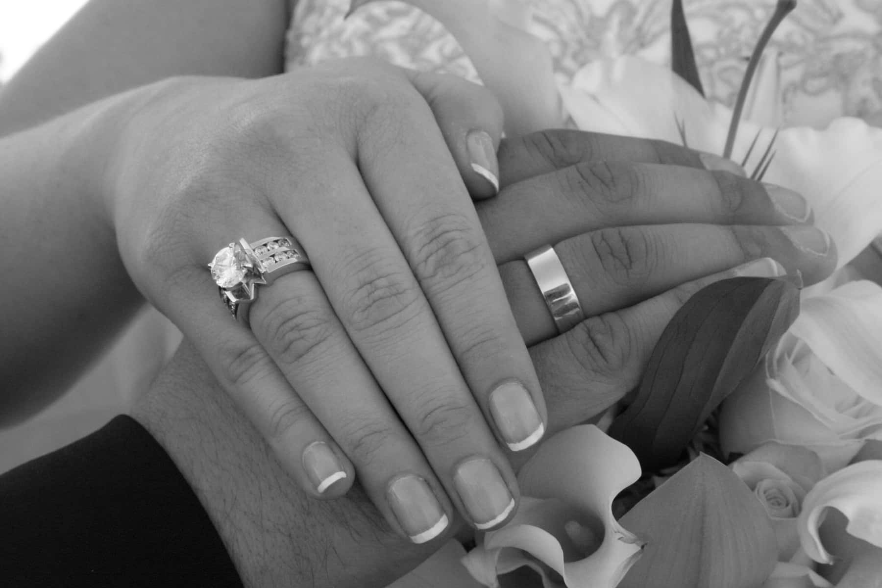兩隻手緊握在一起，戴著結婚戒指，附近有白色花朵。