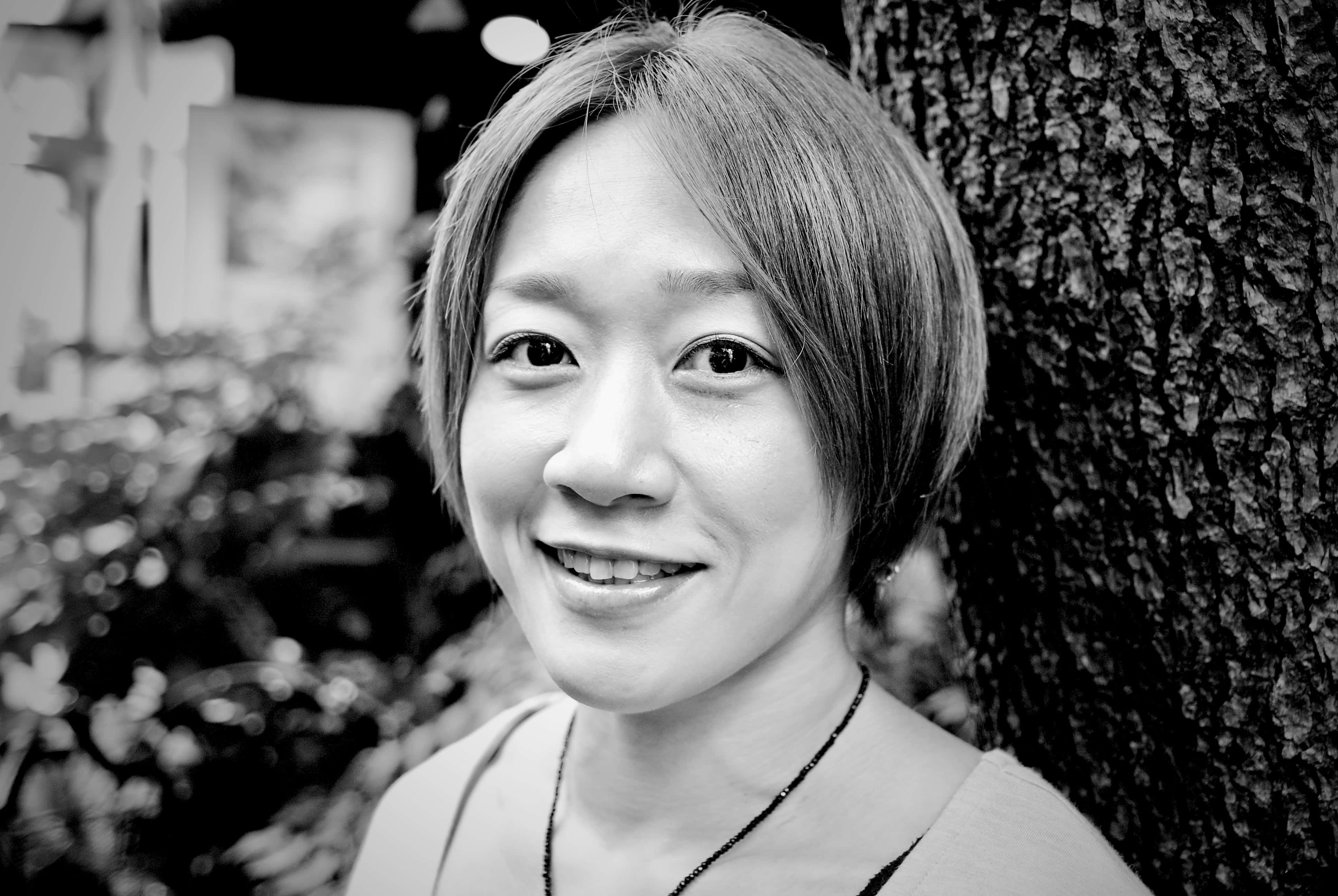 黑白日本綜合格鬥選手梅古美·藤井 壁紙 — 一位短髮微笑的女性靠著紋理豐富的樹幹，柔和的黑白肖像。