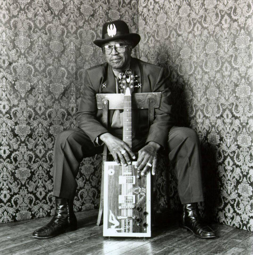 黑白照片：Bo Diddley 坐在椅子上 壁紙 — 一個戴帽子和眼鏡的男人冷靜地坐著，手持一把定制結他，背景是華麗的牆紙，形成一幅黑白肖像。