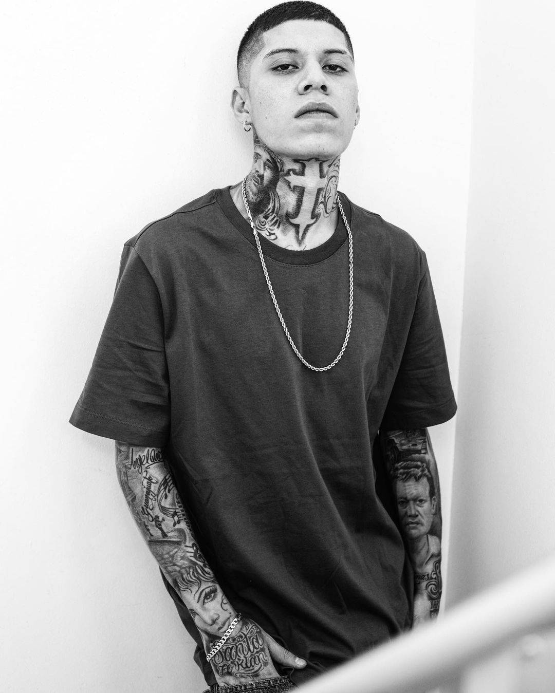 一位紋身男子穿著深色T恤，靠著白牆。