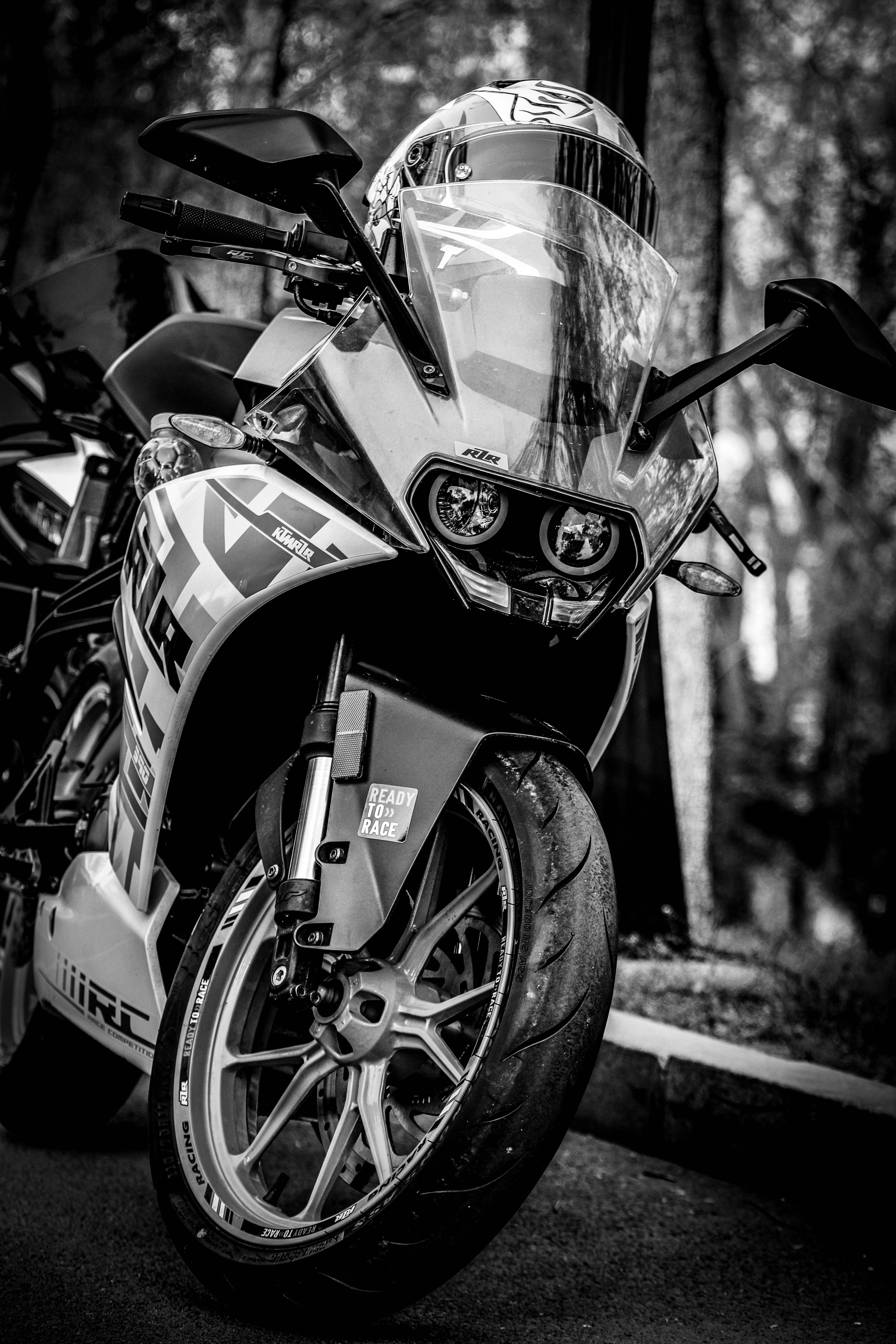 黑白配色KTM RC 390 壁紙 — 一架時尚的黑白運動電單車，頭盔擱在擋風玻璃上，停泊在樹林地區。