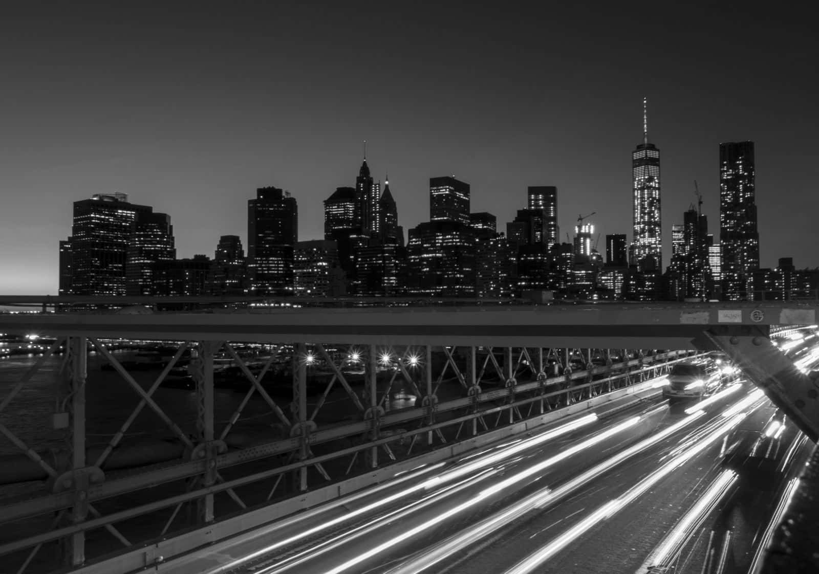 Black And White Cityscape 桌布 壁紙 — 黑白長曝光拍攝的橋樑照片，於黃昏時分，車燈形成光軌，背後是燈火通明的城市天際線。