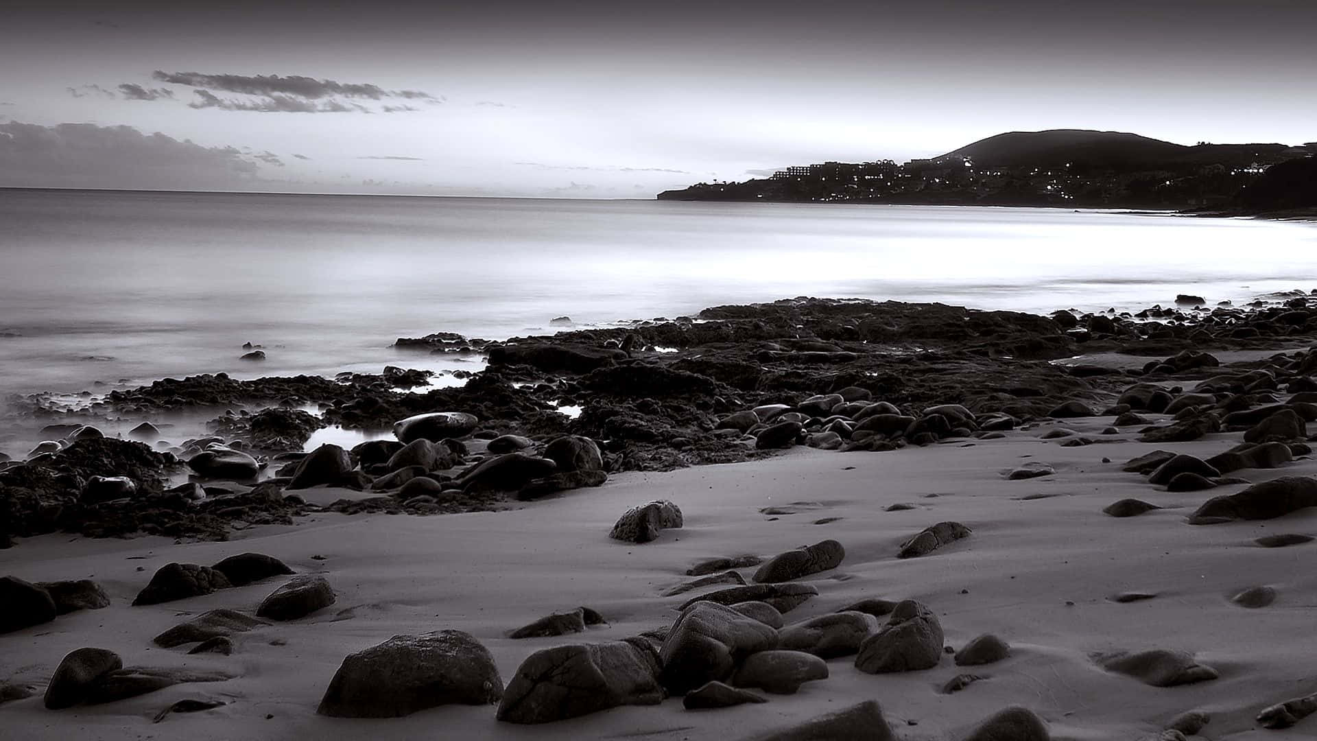 Black And White Landscape 桌布 壁紙 — 黑白長曝光海景，前景有平滑水面和岩石，遠方山城在暮色中發光。
