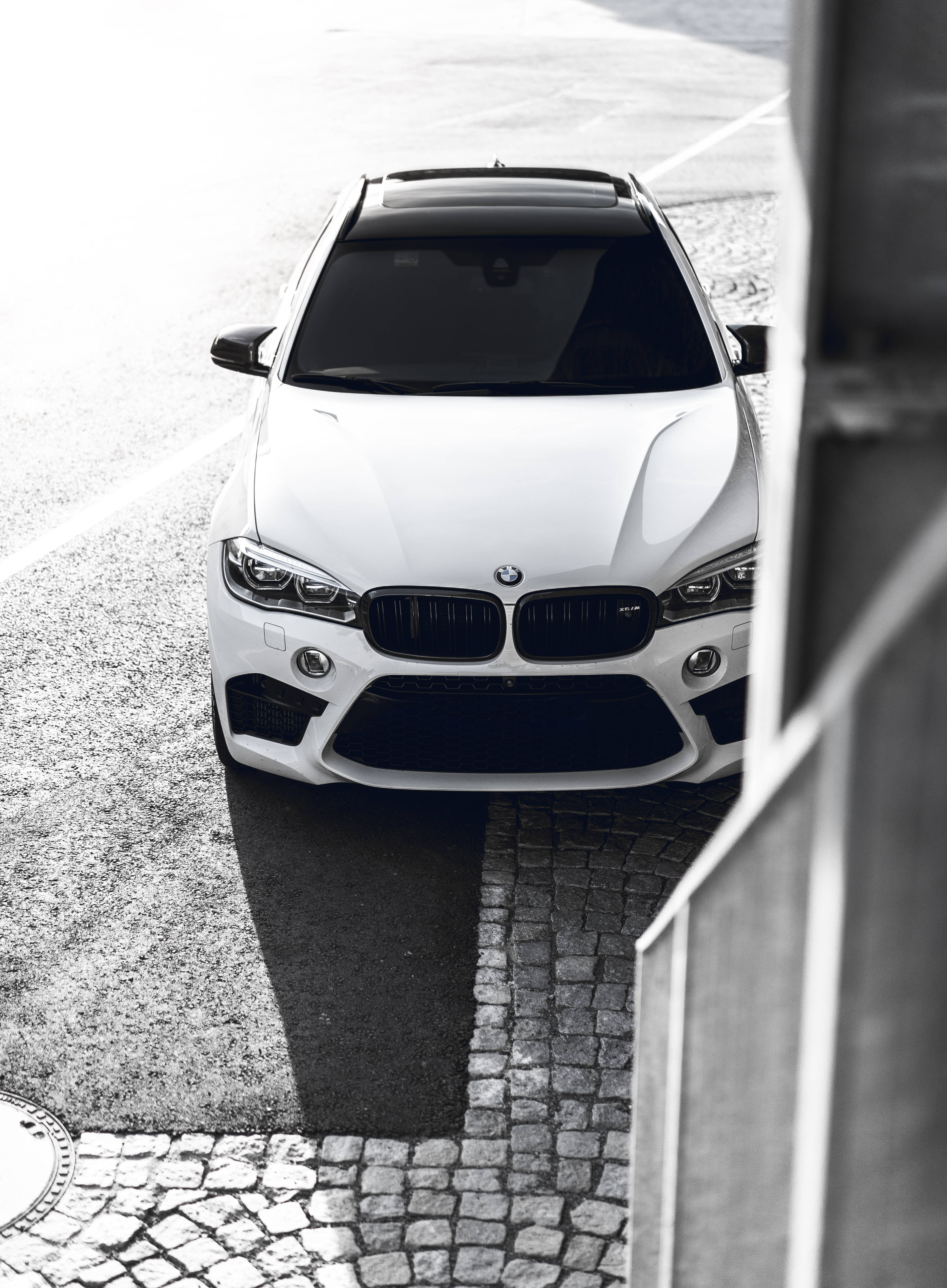 黑白長鏡頭BMW M 壁紙 — 一輛時尚的白色BMW SUV從上方俯瞰在鵝卵石街道上，帶有強烈陰影。
