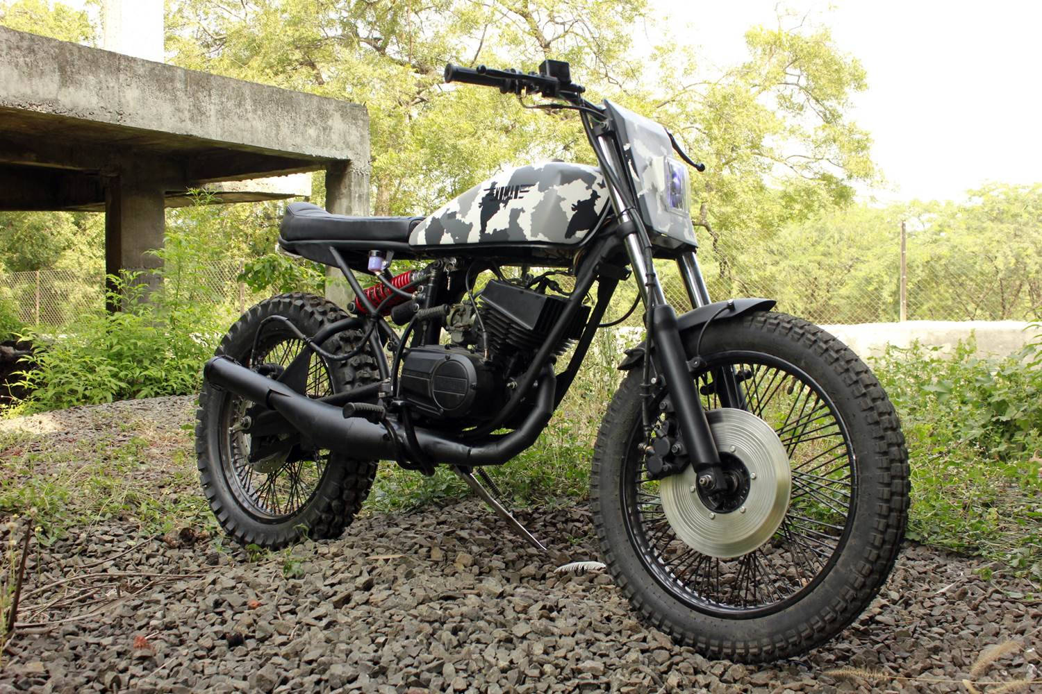 黑白雅馬哈RX100 Scrambler 壁紙 — 一輛定制迷彩圖案的摩托車，配備越野輪胎，停在混凝土高架橋下的礫石上，周圍環繞著綠色植物。