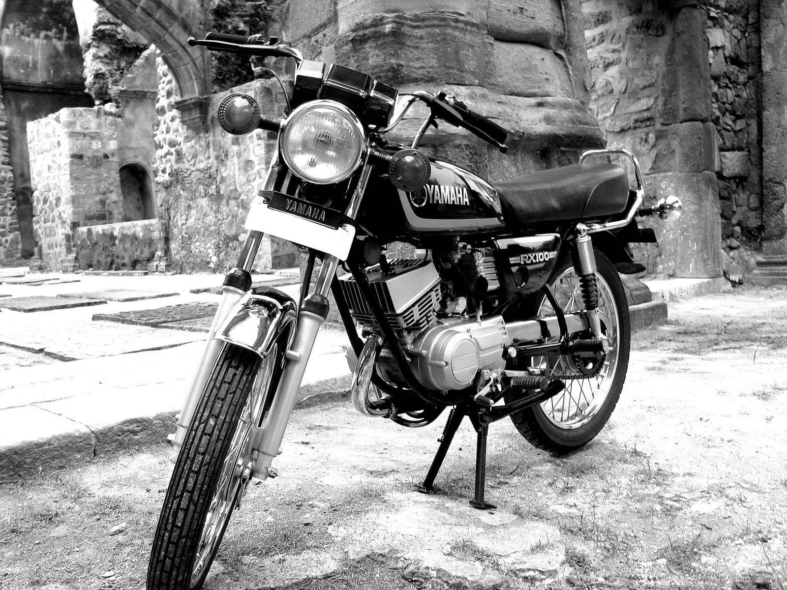 黑白雅馬哈RX100摩托車 壁紙 — 一輛經典的Yamaha FX100電單車以黑白風格矗立在古代廢墟拱門中的石座上。