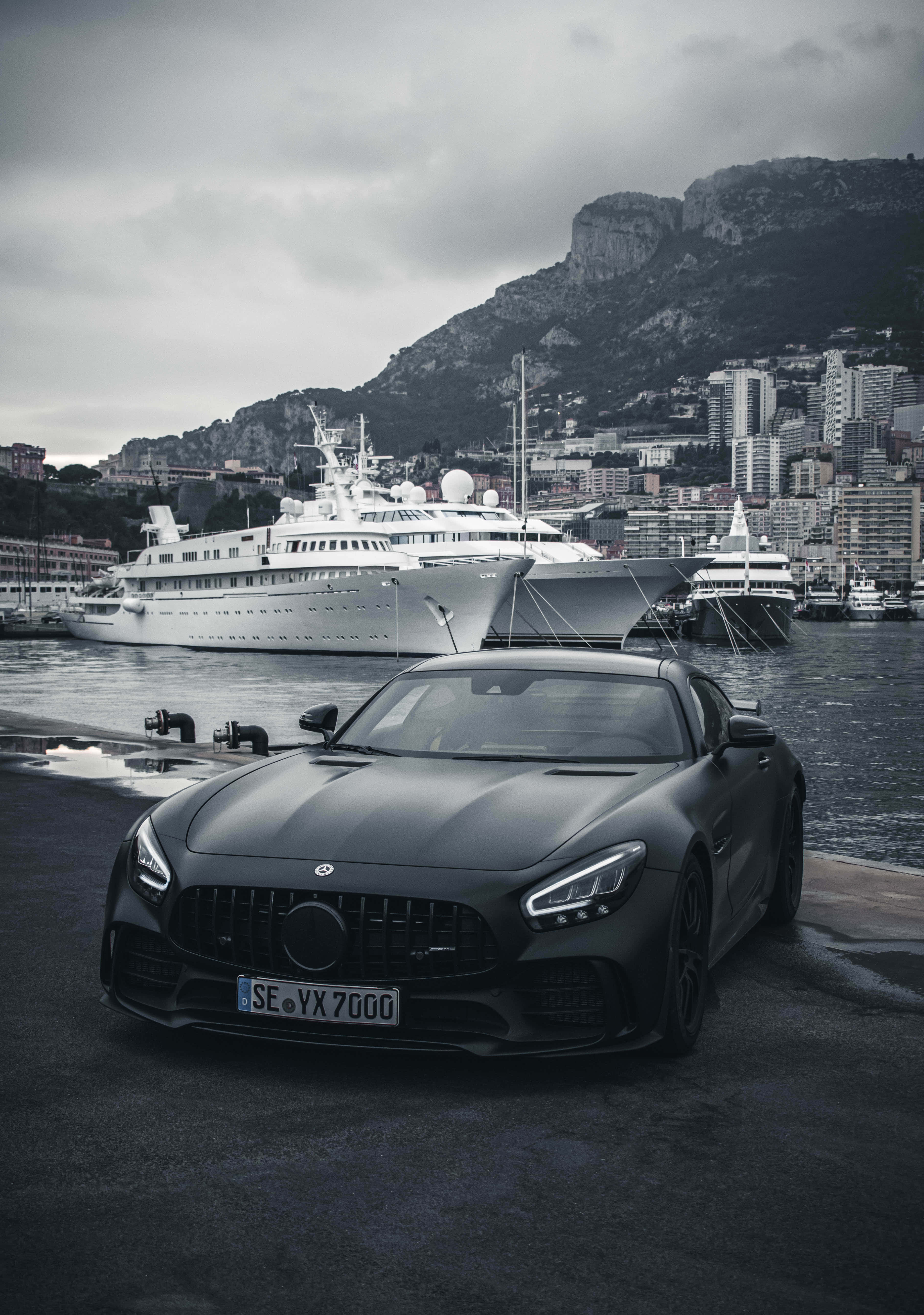 黑白風格AMG GT-R海邊帆船背景壁紙 壁紙 — 一架啞光黑色跑車停泊在港口碼頭，背景有豪華遊艇和陰天下的山區沿海城市。