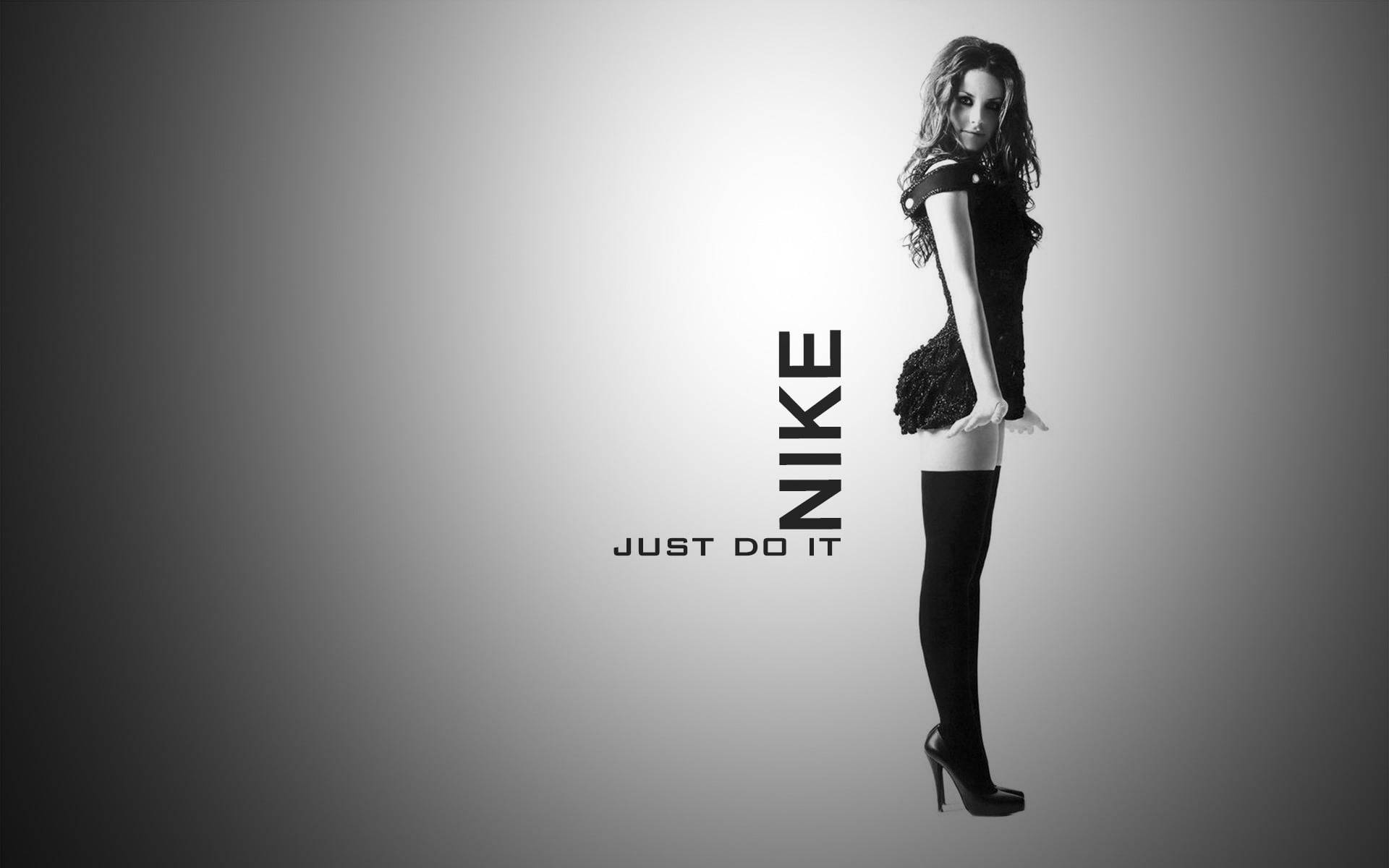 黑白風格的「Just Do It」女孩 壁紙 — 一位穿著高跟鞋和短裙的女性在Nike Just Do It標誌旁擺姿勢的黑白工作室肖像。