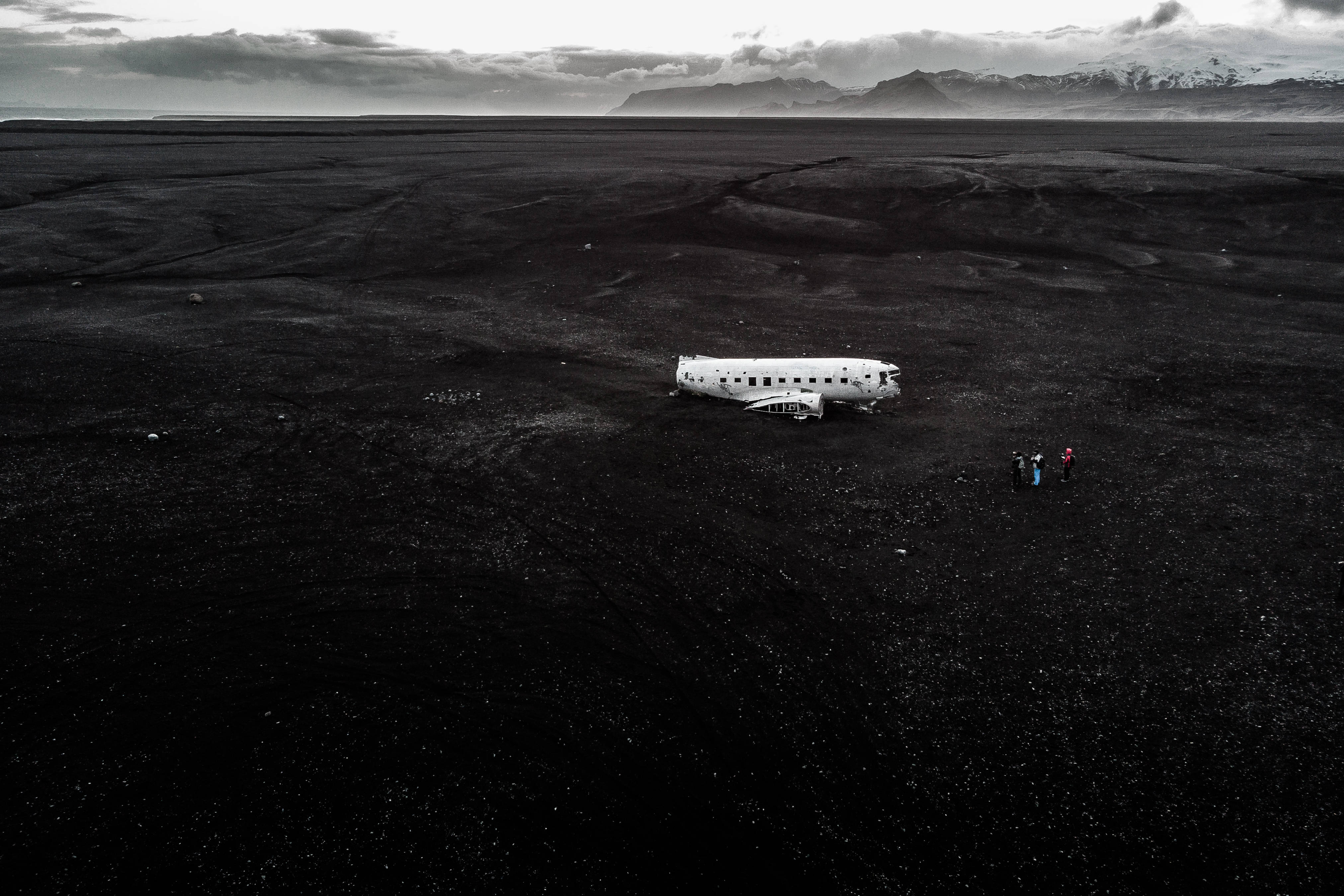 黑白高清墜毀飛機 壁紙 — 一架孤獨的白色飛機機身停放在廣闊的黑色火山岩場上，遠方有積雪的山峰，天空多雲。