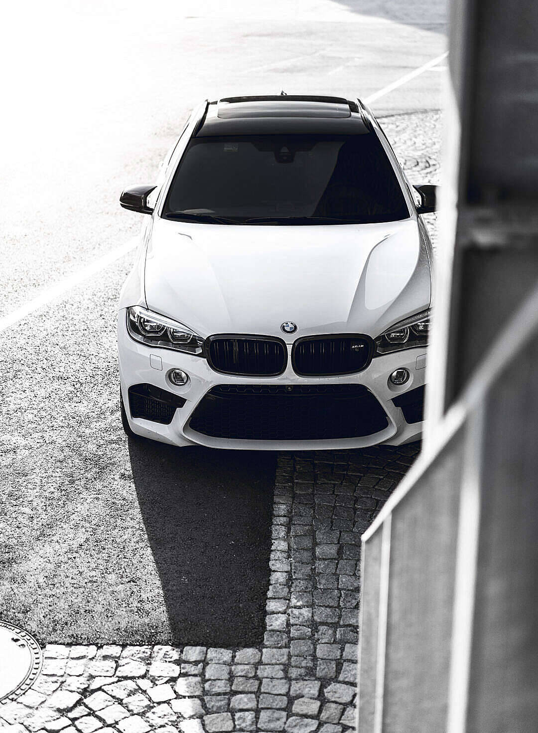 黑白高清汽車 壁紙 — 一架流線型白色BMW SUV從上方拍攝，停泊在鵝卵石街道上，強烈的陰影和城市建築構成了畫面的框架。