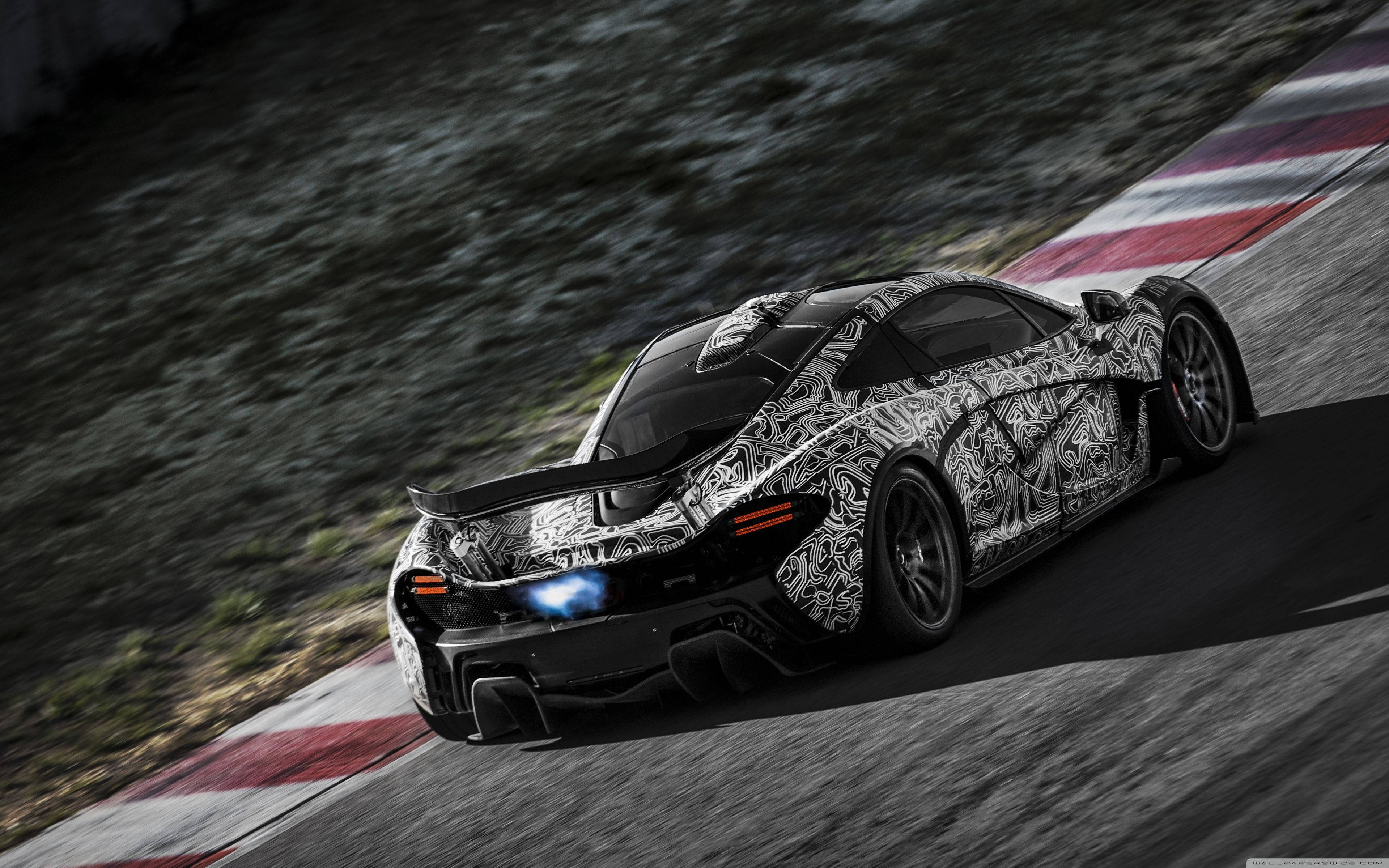 黑白麥拉倫P1 壁紙 — 一輛迷彩McLaren超級跑車在明亮的日光下沿著彎曲的賽道奔馳，賽道有紅色和白色的路緣。