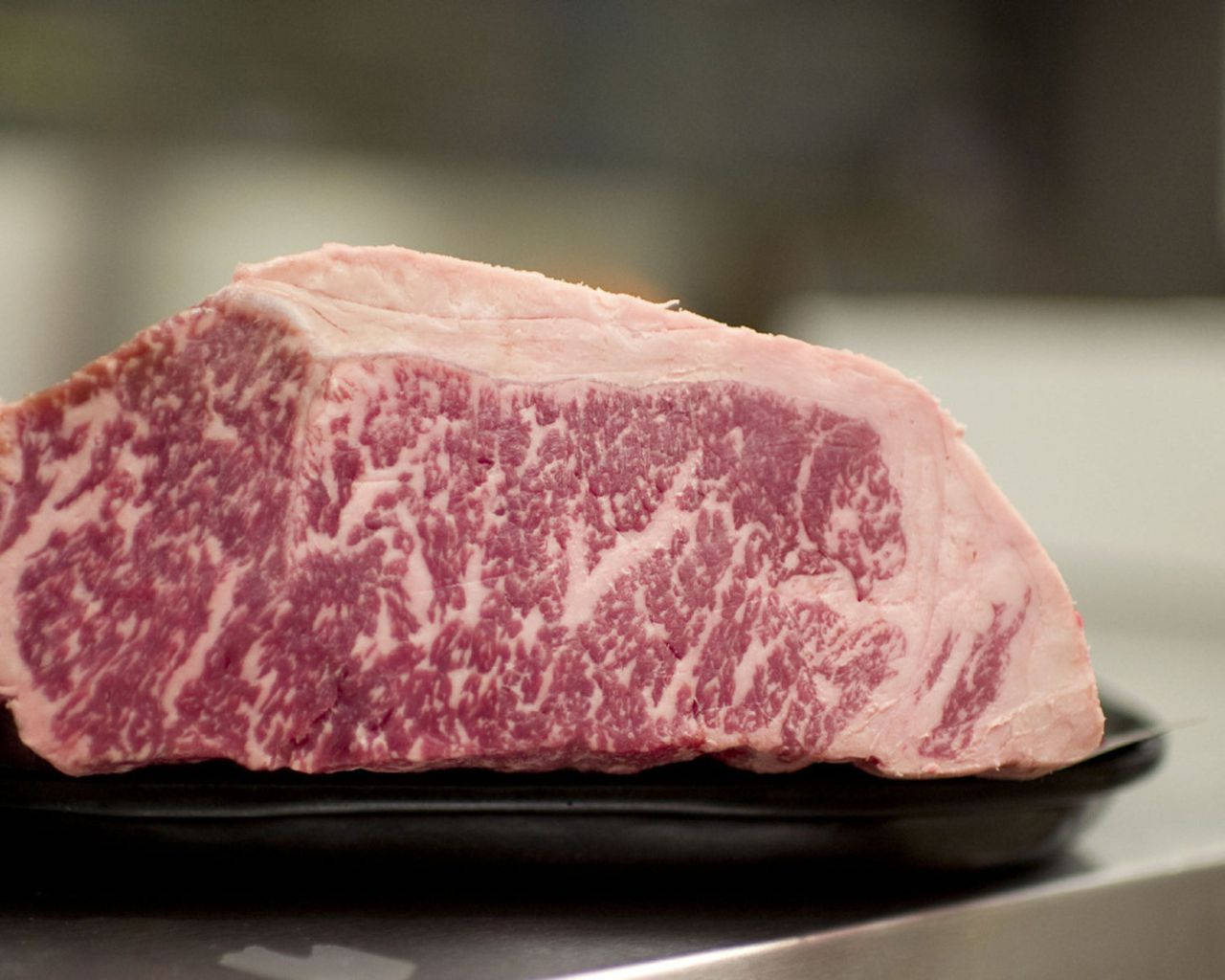 在廚房環境中，黑色碟上放著一塊生的大理石紋牛肉扒。